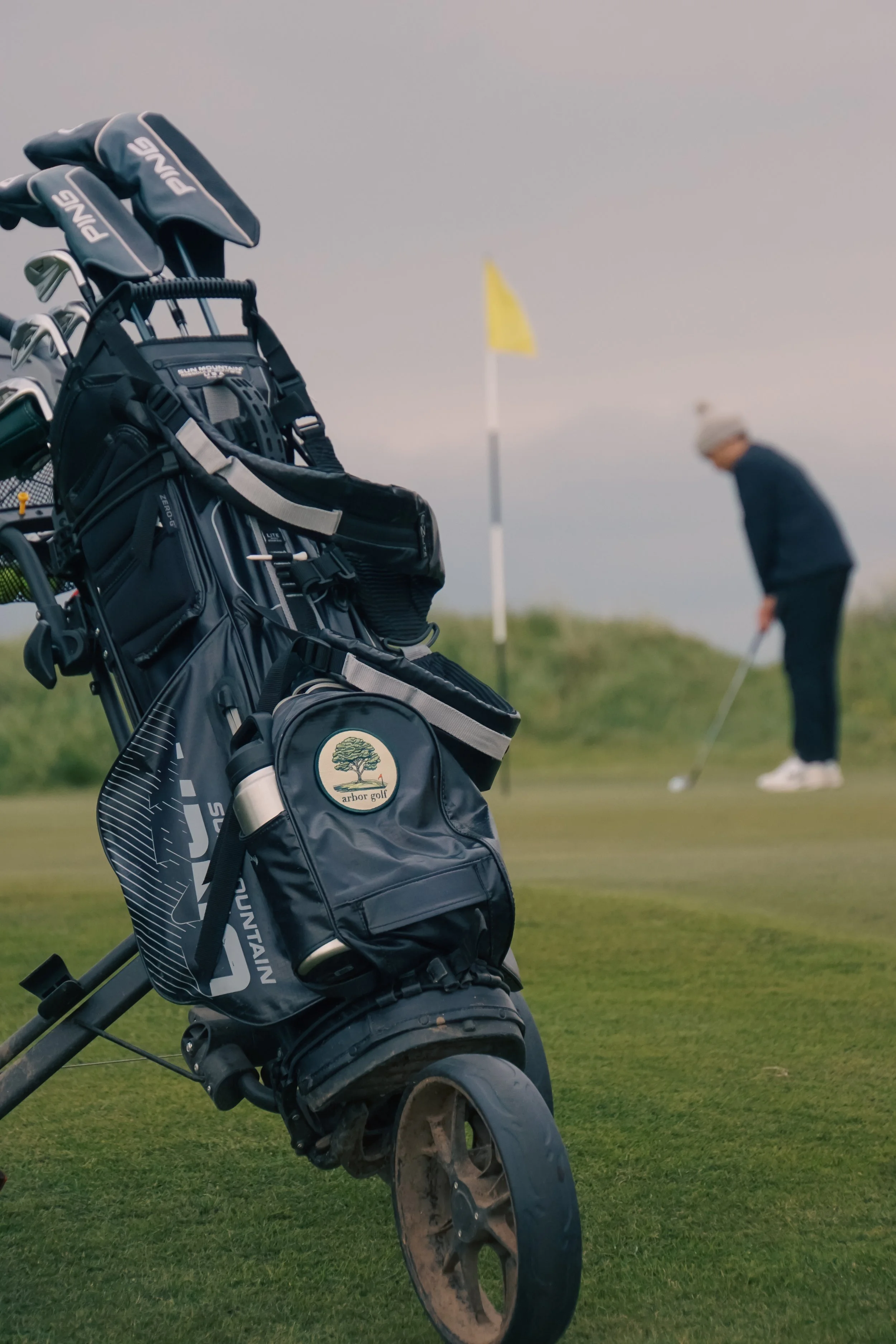 Golf bag on a pushcart with a golfer preparing to putt in the background near a flag on a green.