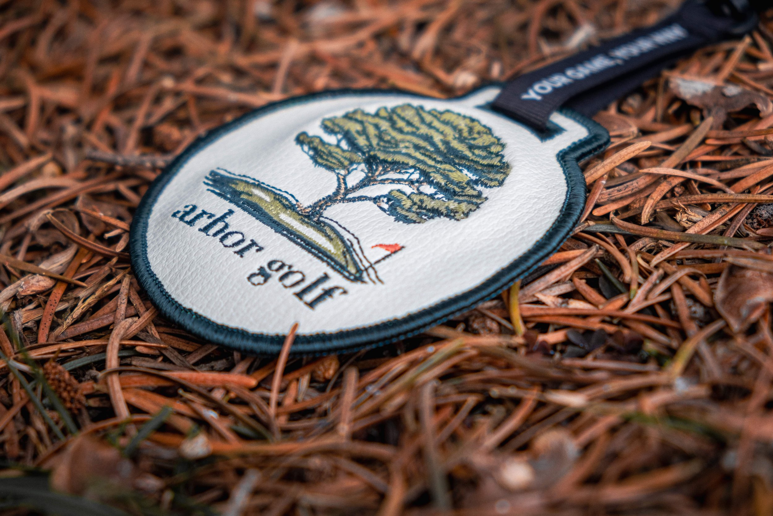 Golf-themed leather tag with tree logo and "arbor golf" text on pine needles.