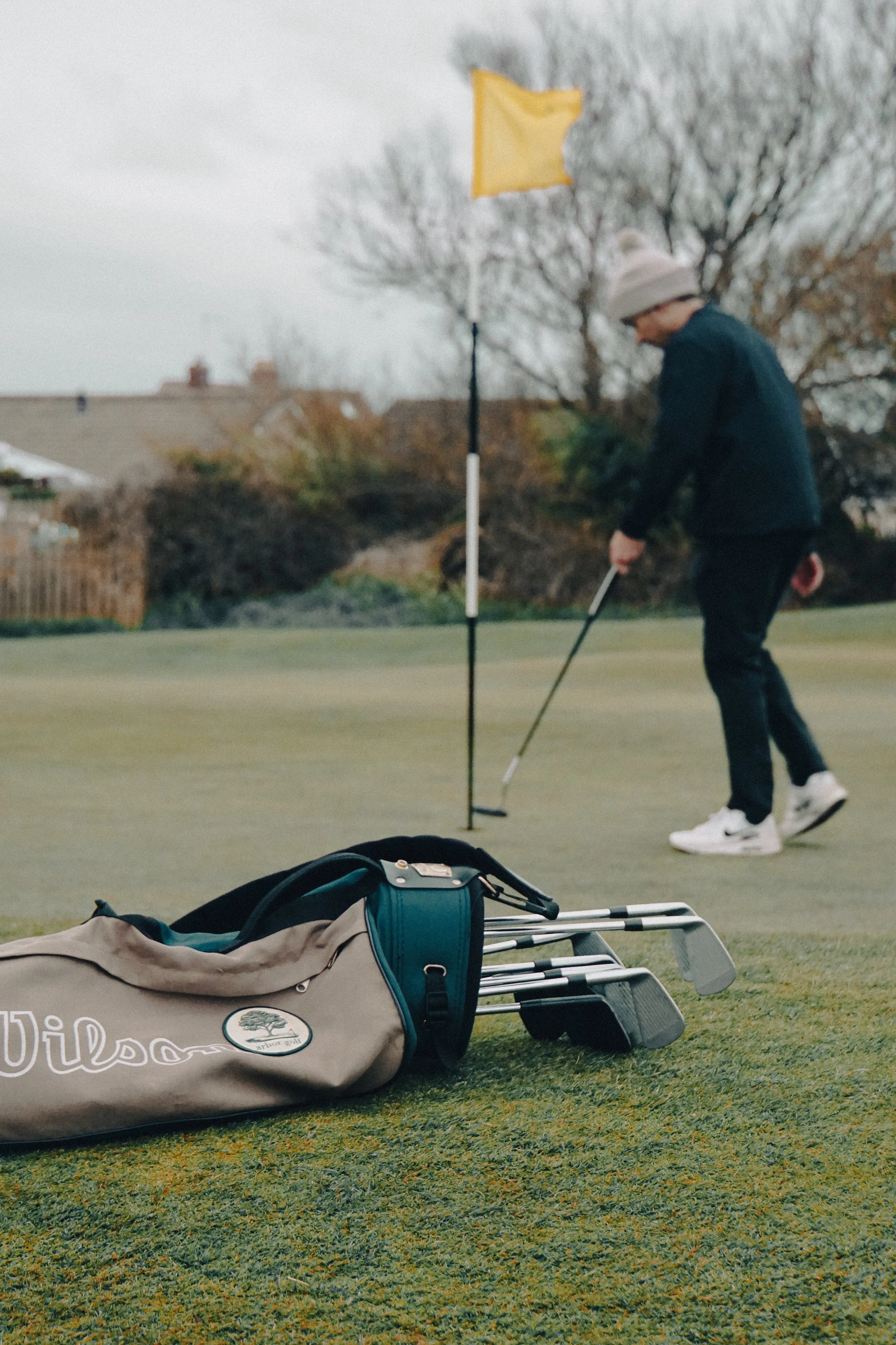 Golfer putting on a green near flag, golf bag with clubs in foreground, overcast weather.