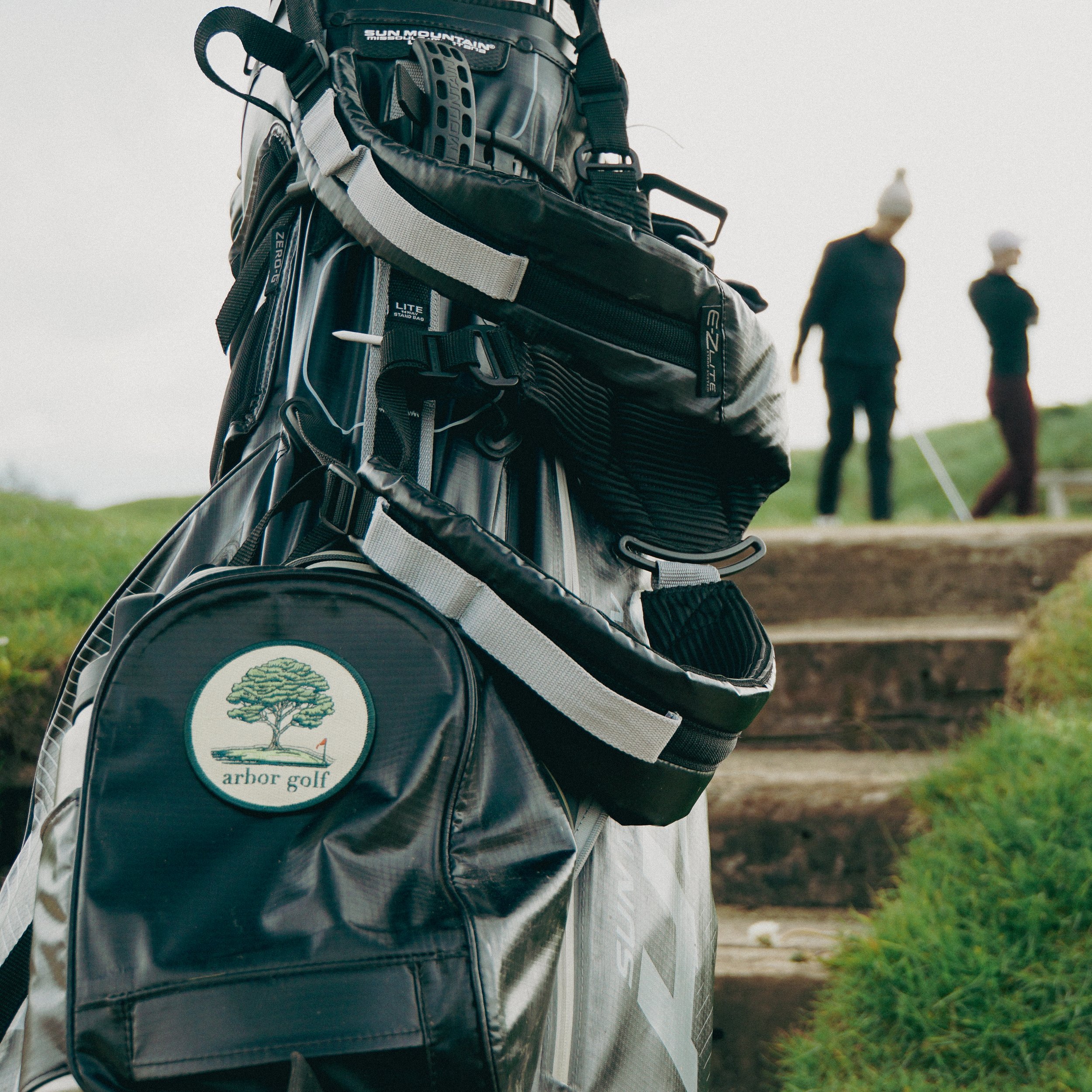 Close-up of a black golf bag with a patch labeled "Arbor Golf" featuring a tree, with two blurred figures in the background on a golf course.