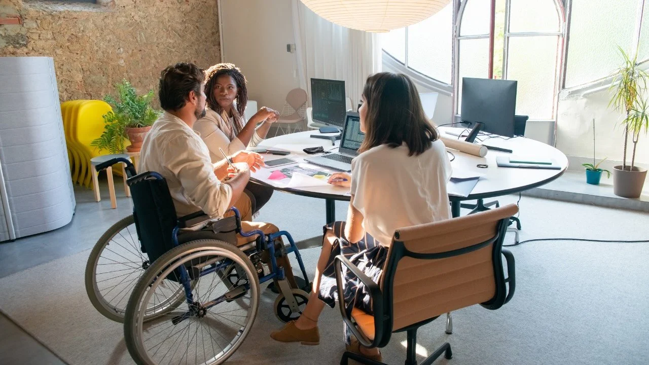 Team sitting around a circular table with a laptop, computer screens, and colorful sticky notes.