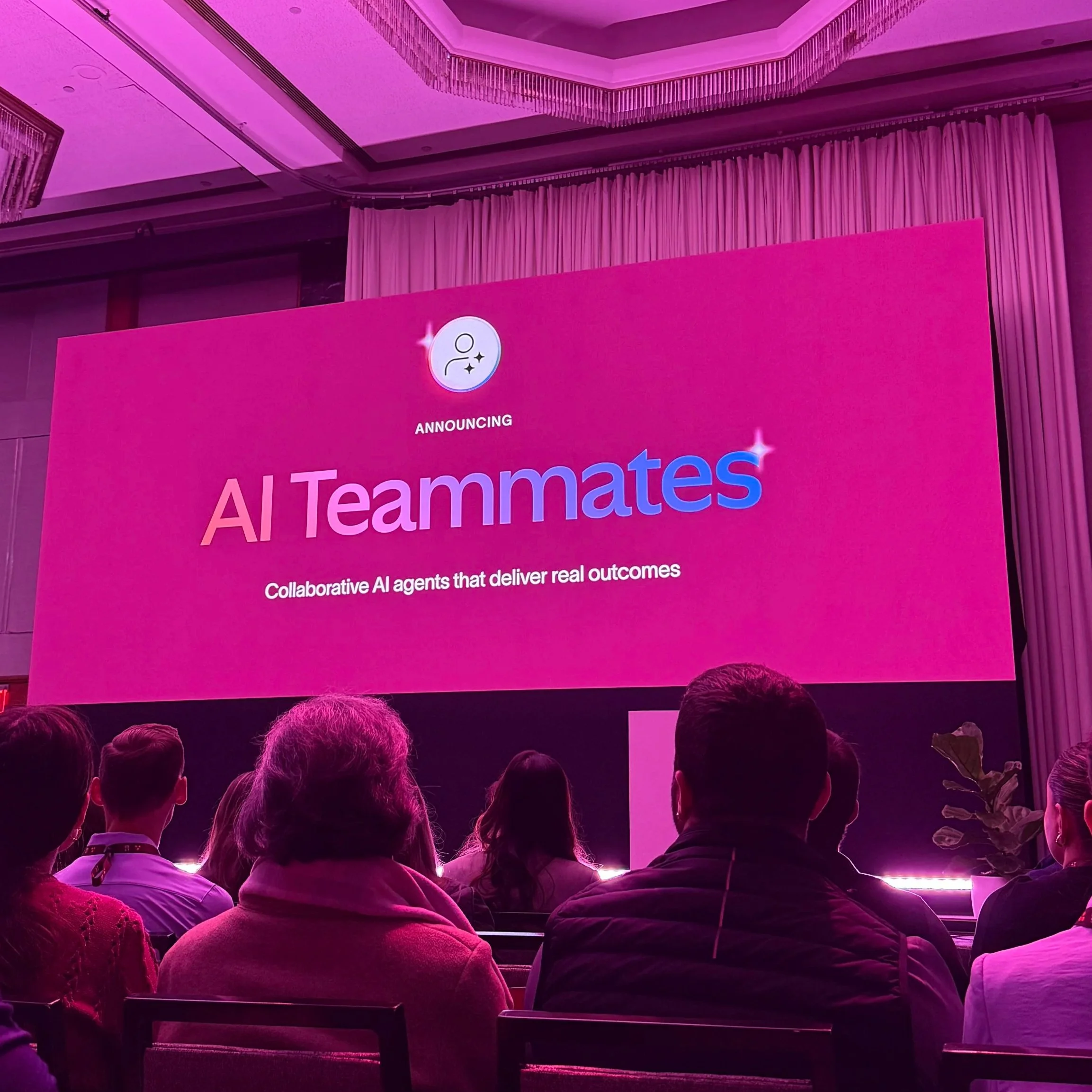A room full of people watch a presentation on Asana AI Teammates. There is a large screen that displays the text "Announcing AI Teammates. Collaborative AI agents that deliver real outcomes."