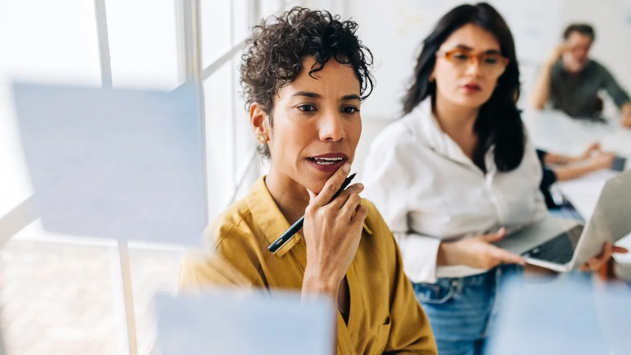 A woman in the foreground looking at a clear panel with white note paper hanging on it, two teammates in the background working with laptops and papers.