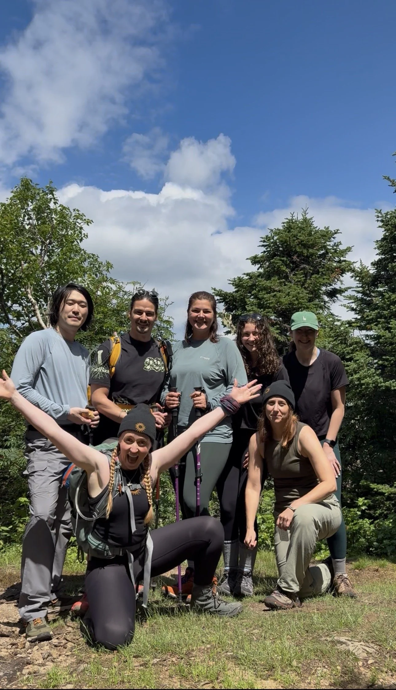 Group of seven hikers in outdoor nature setting, surrounded by green trees, with a bright blue sky and some clouds overhead.