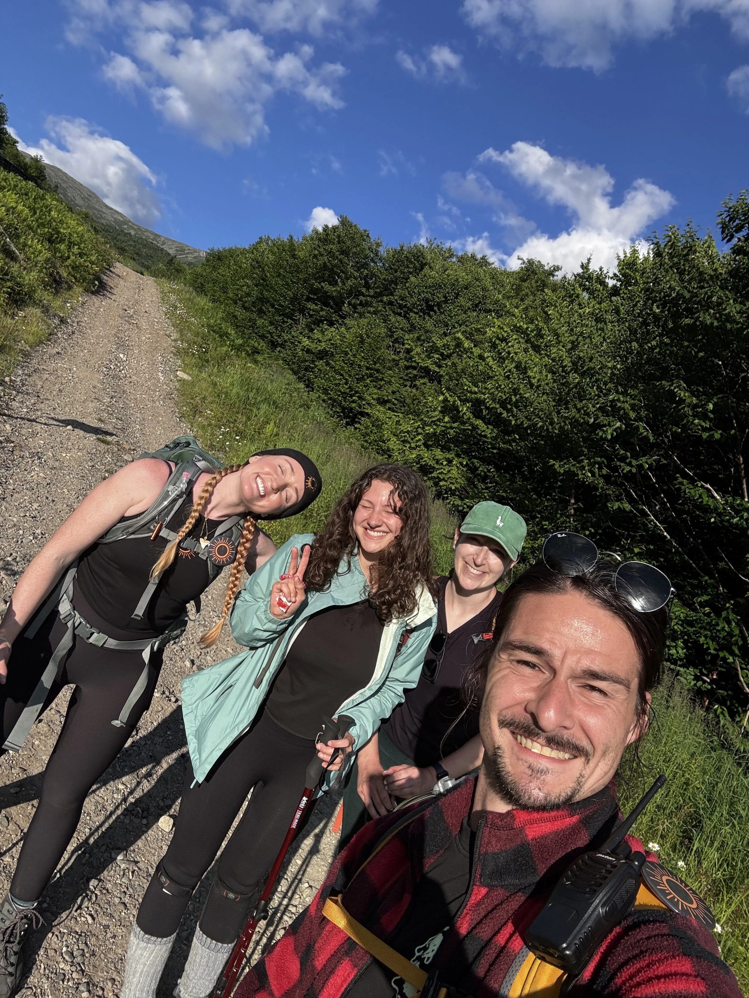 Group of five smiling hikers taking a selfie on a mountain trail with green trees and a blue sky with clouds in the background.