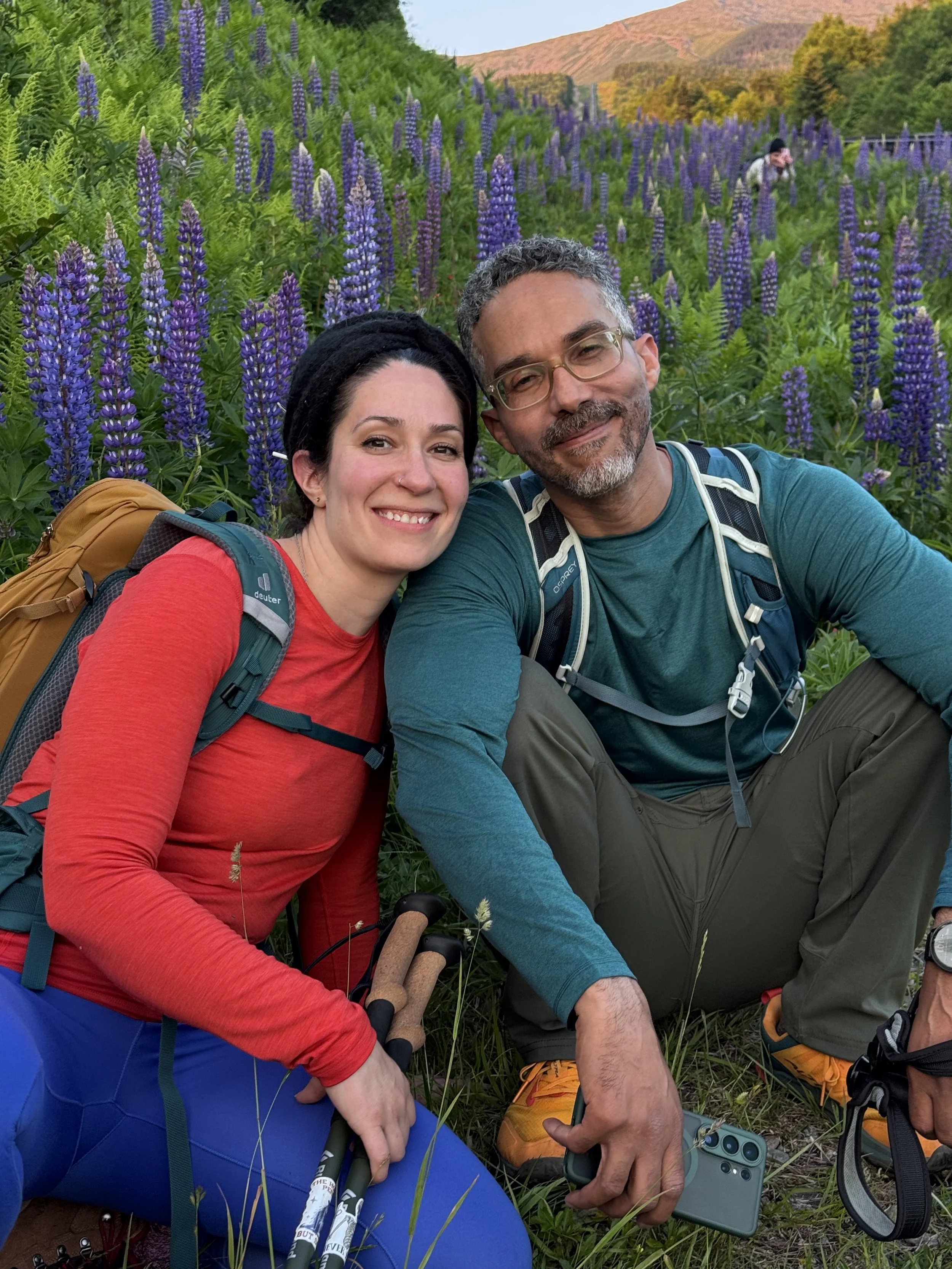 A smiling woman and man sitting on the grass in a field of purple flowers, with trees and hills in the background. They are wearing outdoor gear, backpacks, and holding trekking poles, taking a selfie with a smartphone.