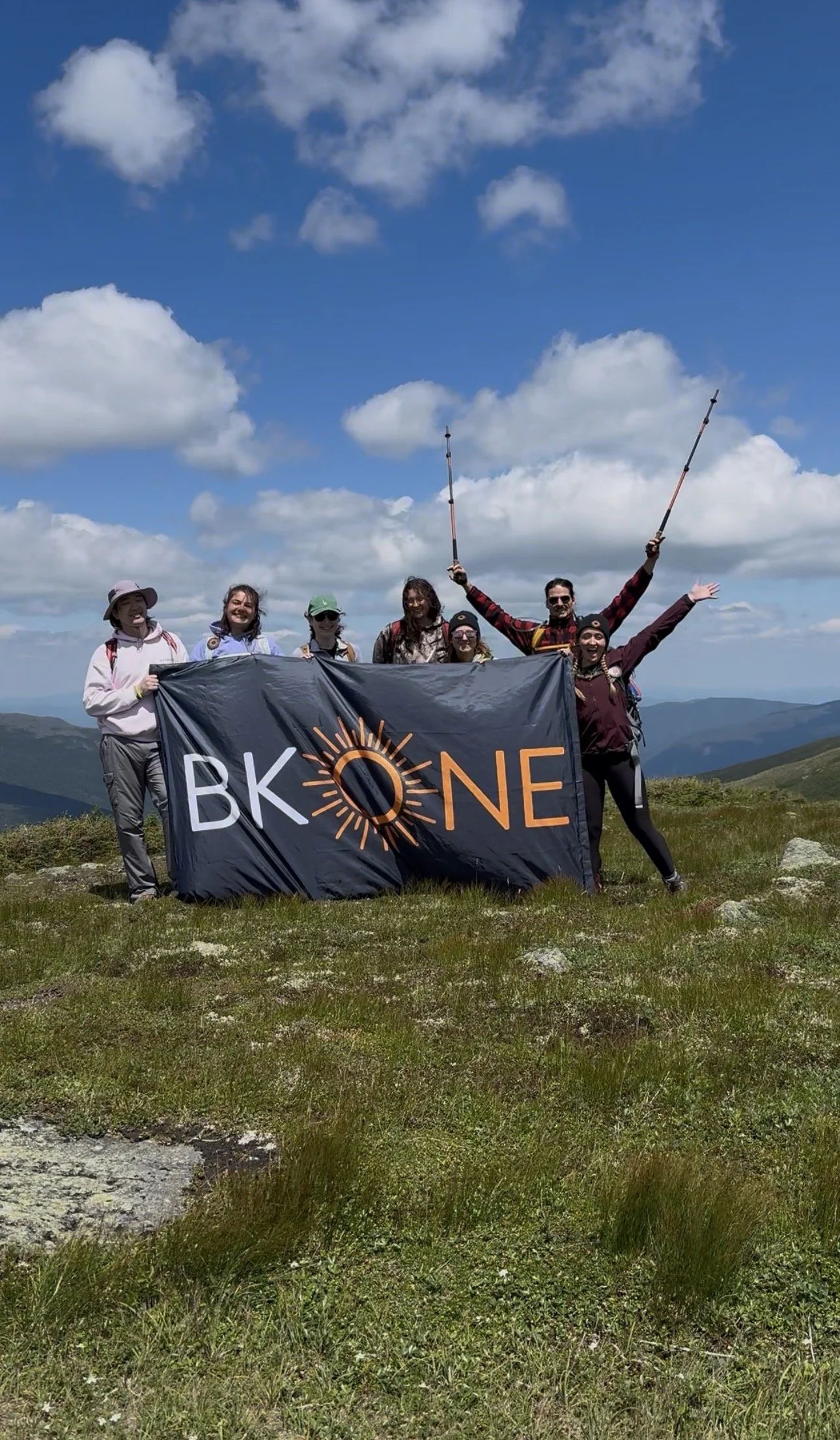 BK:ONE community holding the studio flag on a grassy summit during the Empowered On Earth retreat in the White Mountains.