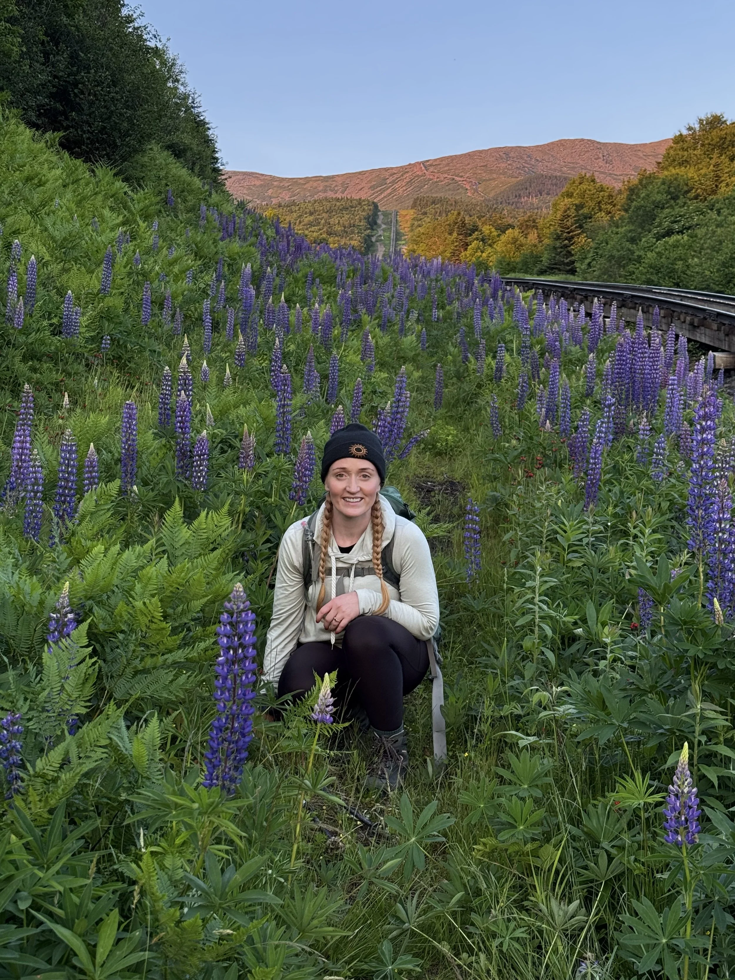 A woman with long braided hair and a black beanie smiling and crouching in a field of tall purple flowers with green foliage, a train track nearby, and a mountain with sparse trees in the background under a clear sky.
