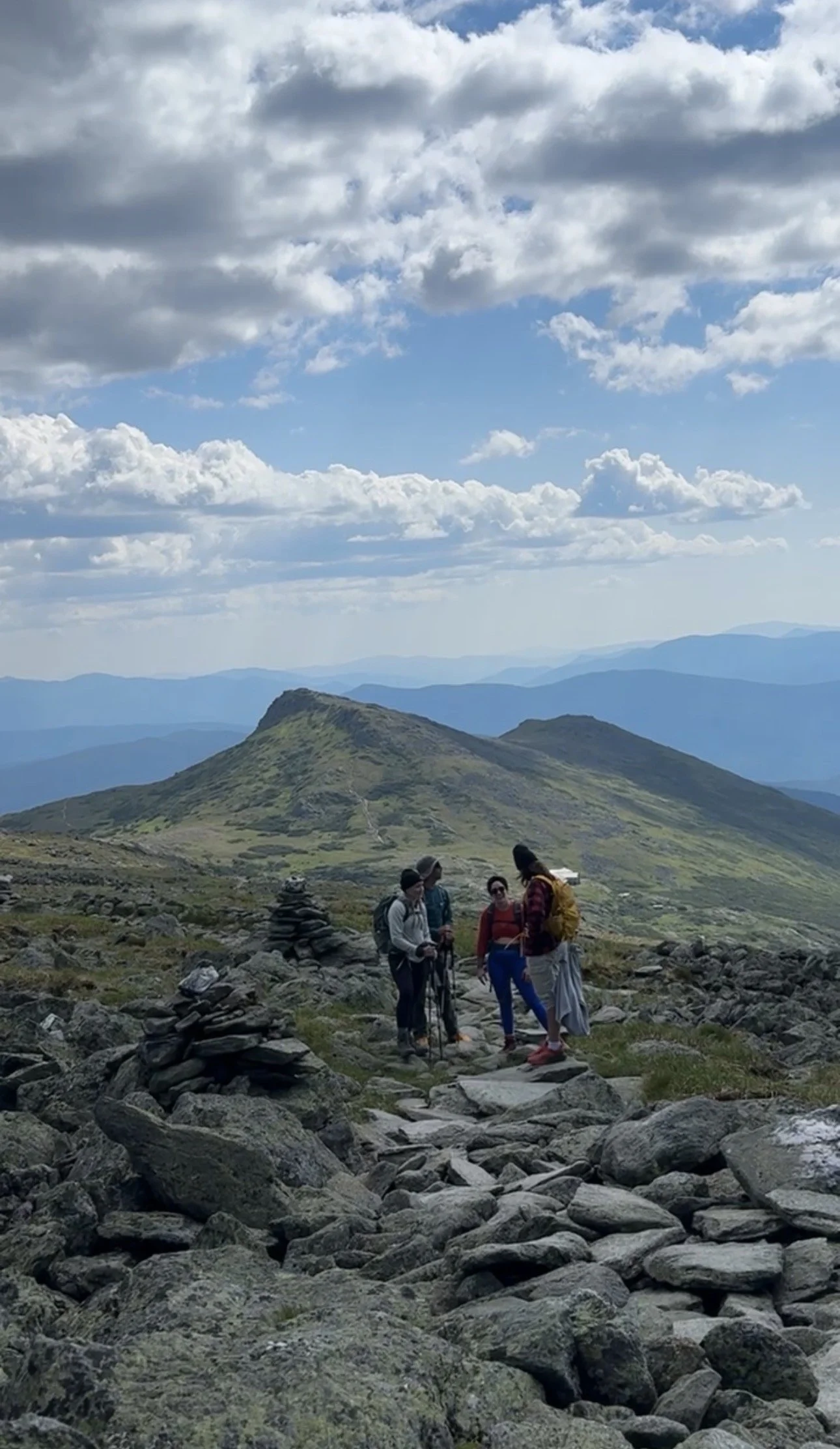 A group of hikers standing on a rocky trail in a mountainous landscape with cloudy sky and distant mountain ridges.