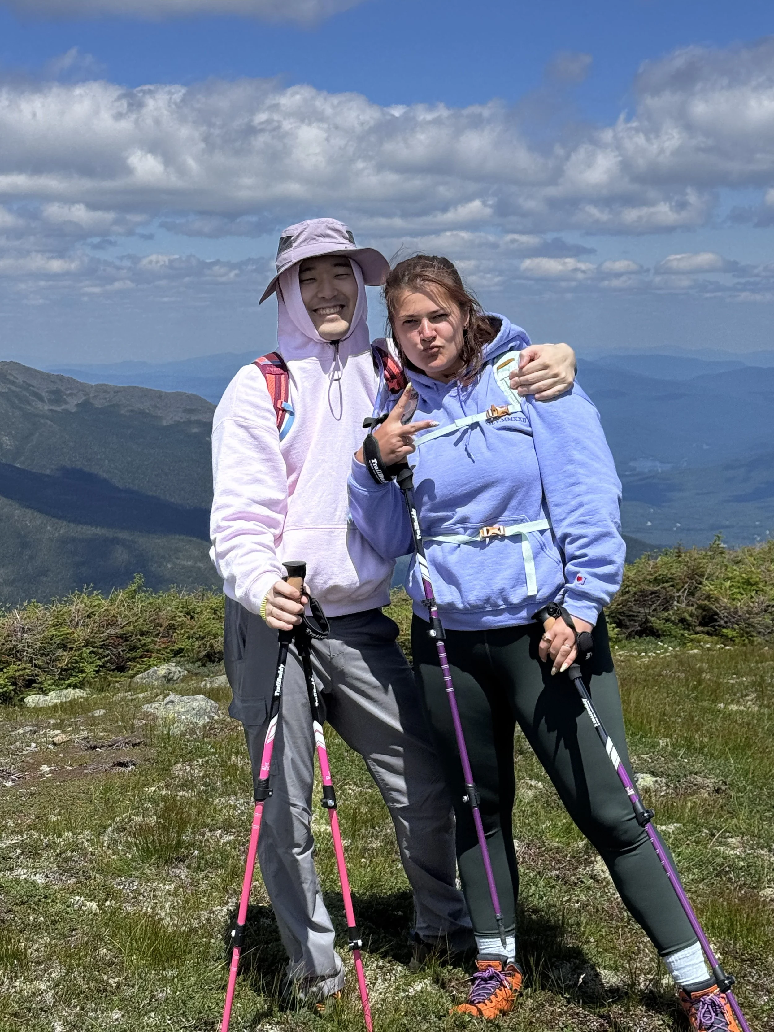 Two smiling women with backpacks and hiking poles standing on a mountain with a scenic view of rolling hills and a partly cloudy sky.