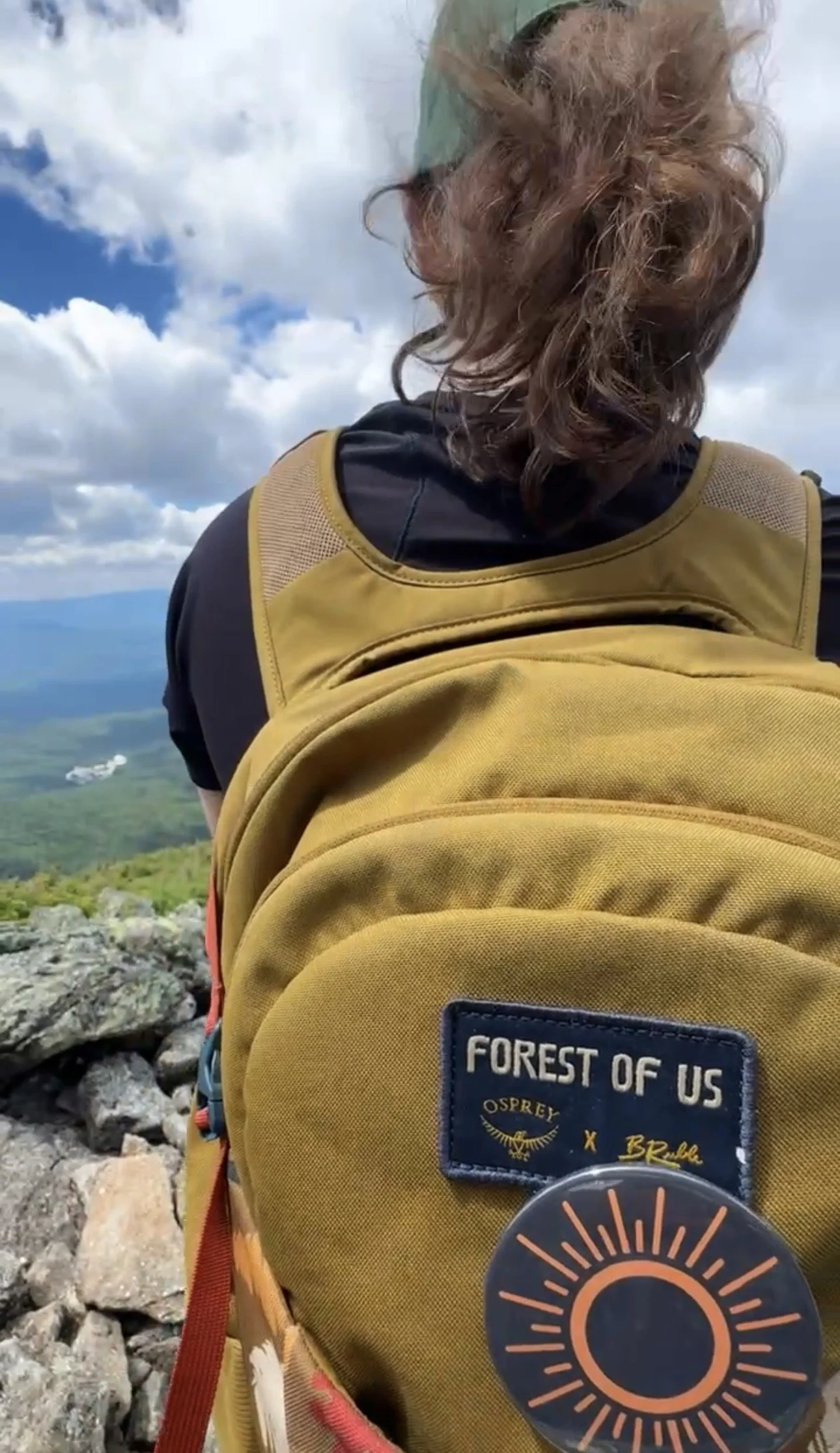 A person with long, curly hair wearing a black and yellow hiking backpack with a patch reading 'Forest of US' and a circular design sticker, sitting on a rocky mountain trail, overlooking a scenic view of rolling hills and a sky filled with clouds.