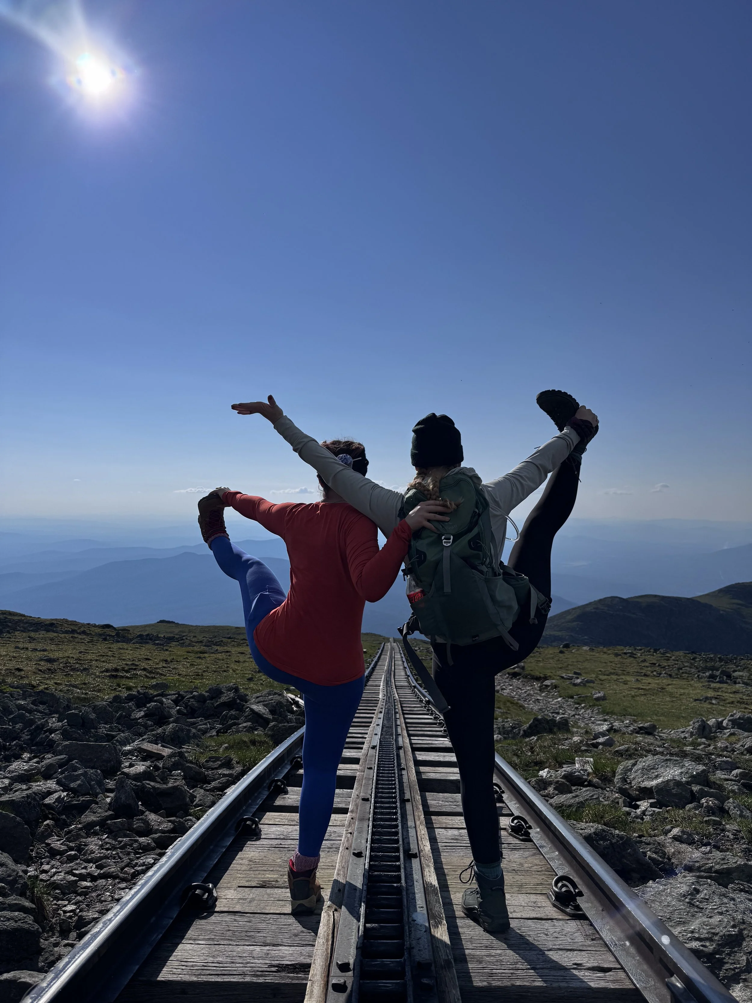 Two people in athletic clothing and backpacks are doing a yoga pose on a railroad track in a mountain landscape. They are holding each other's arms and lifting one leg each, with arms outstretched.