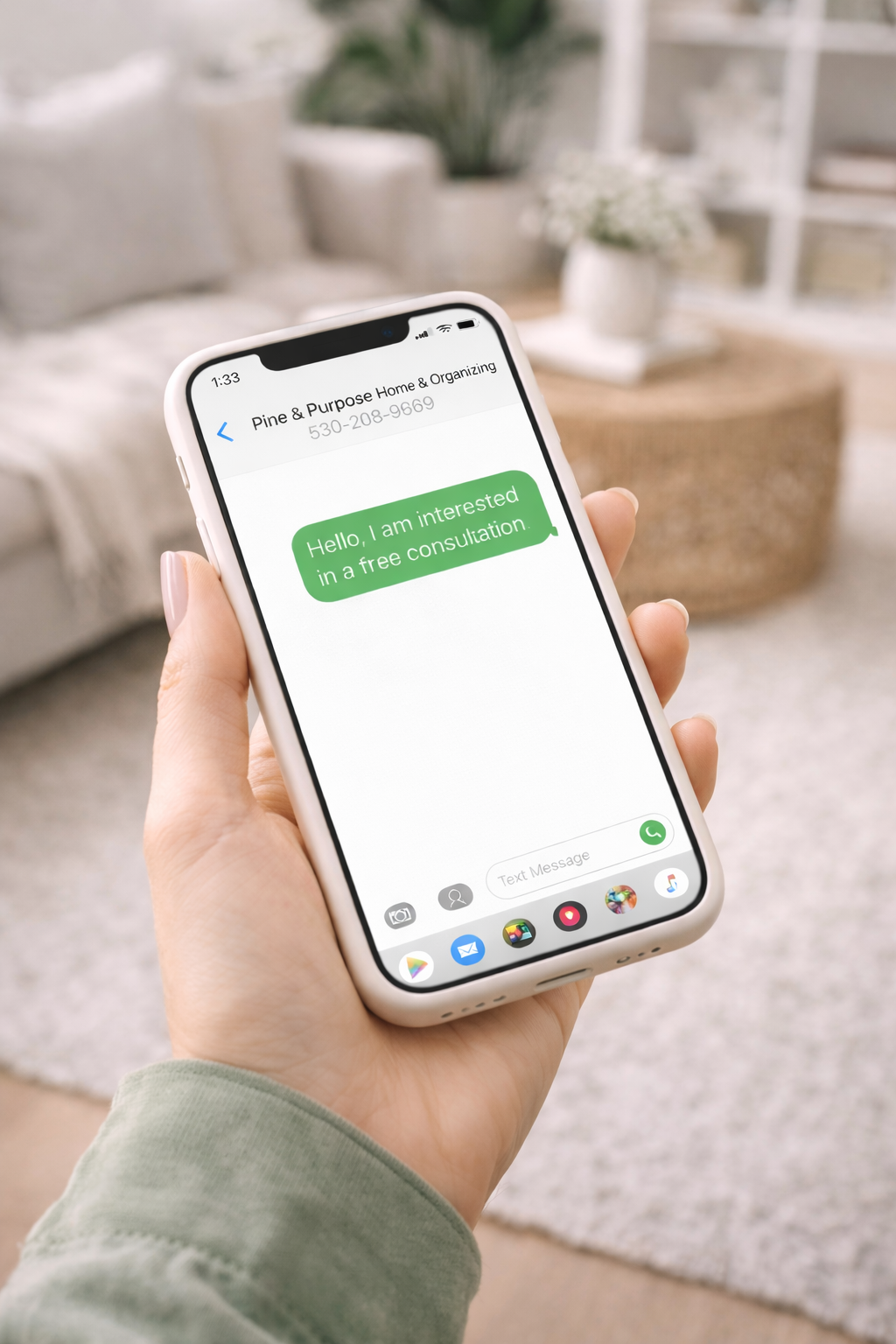 A person holding a smartphone displays a text message conversation with 'Pine & Purpose Home & Organizing' where the person has sent a message saying 'Hello, I am interested in a free consultation'. The background shows a cozy, well-decorated living room with a beige sofa, a round wicker coffee table, a gray rug, and some plants and books in the background.