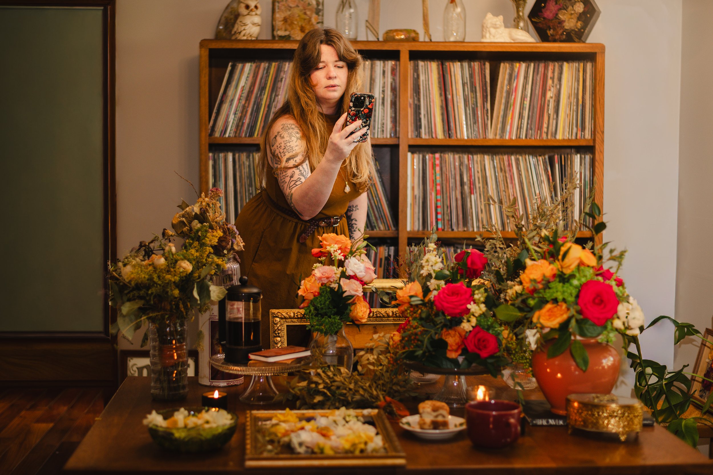 A woman with long hair and tattoos on her arms holding a smartphone, standing behind a table decorated with colorful flower arrangements and candles, in a room with a wooden bookshelf filled with vinyl records.