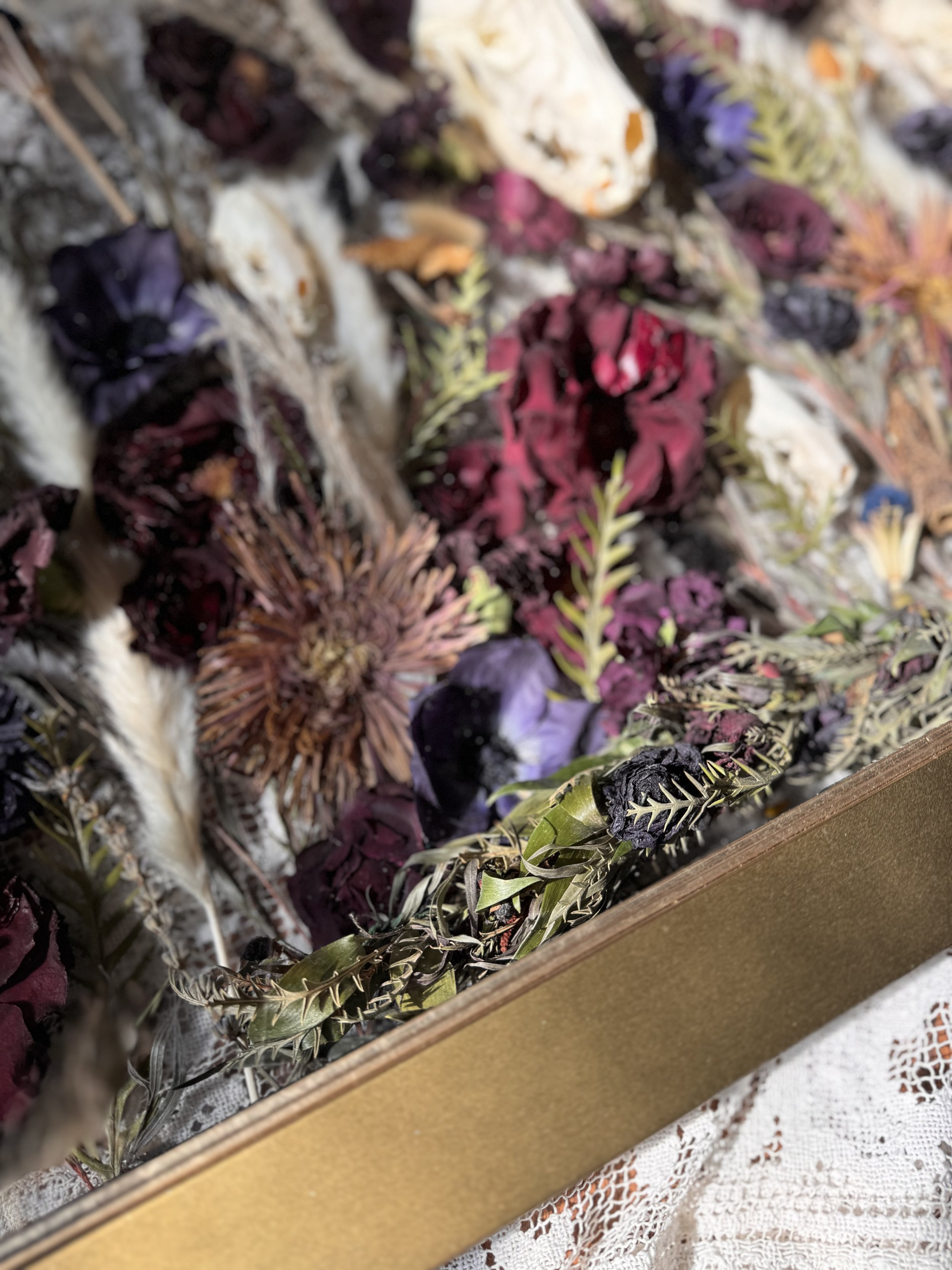 Close-up of a box filled with dried flowers, including roses and other assorted types, alongside dried foliage and grasses on a lace tablecloth.