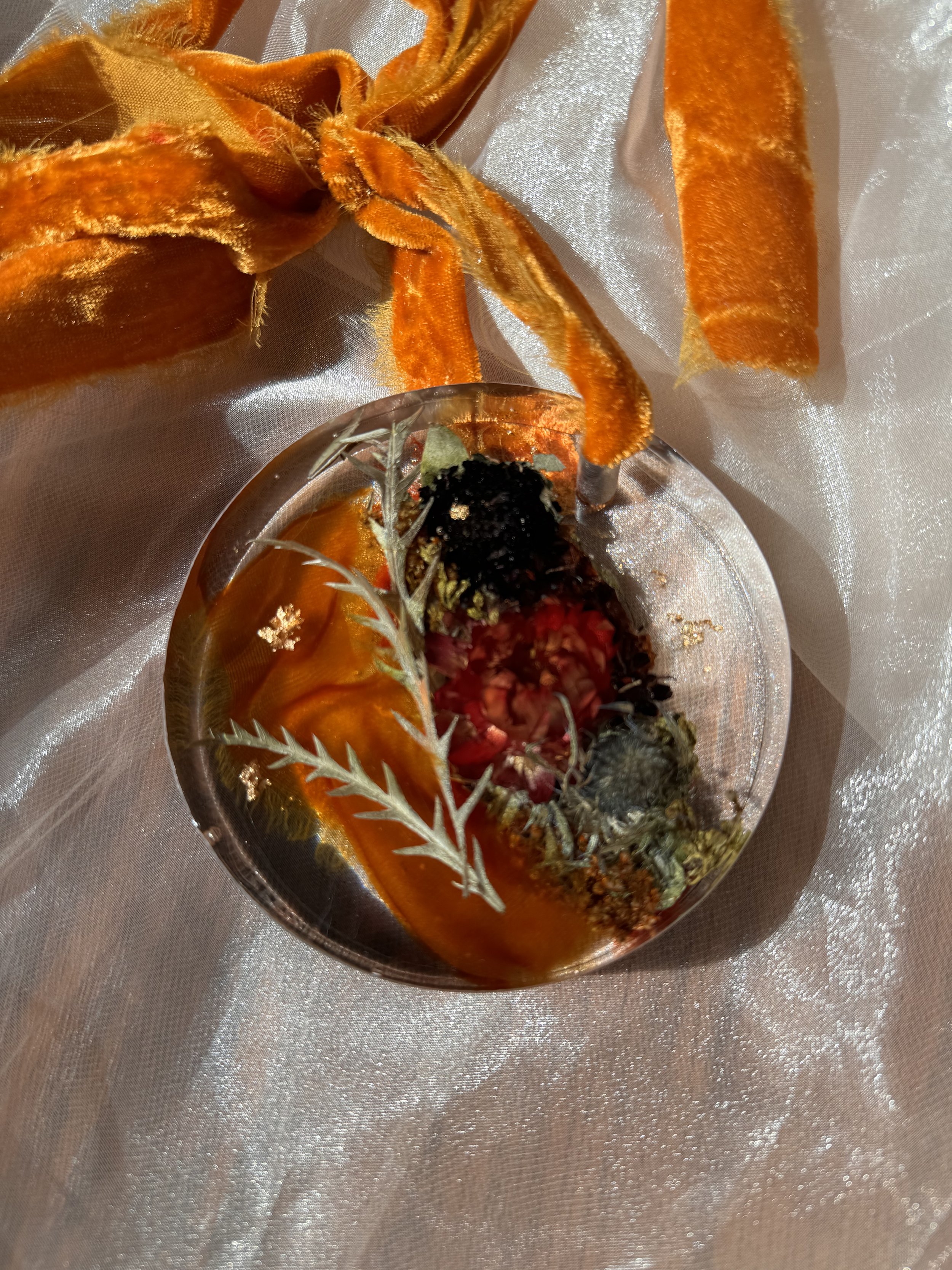 A decorative pendant with dried flowers and leaves encased in resin, accompanied by an orange velvet ribbon, placed on a silky fabric background.
