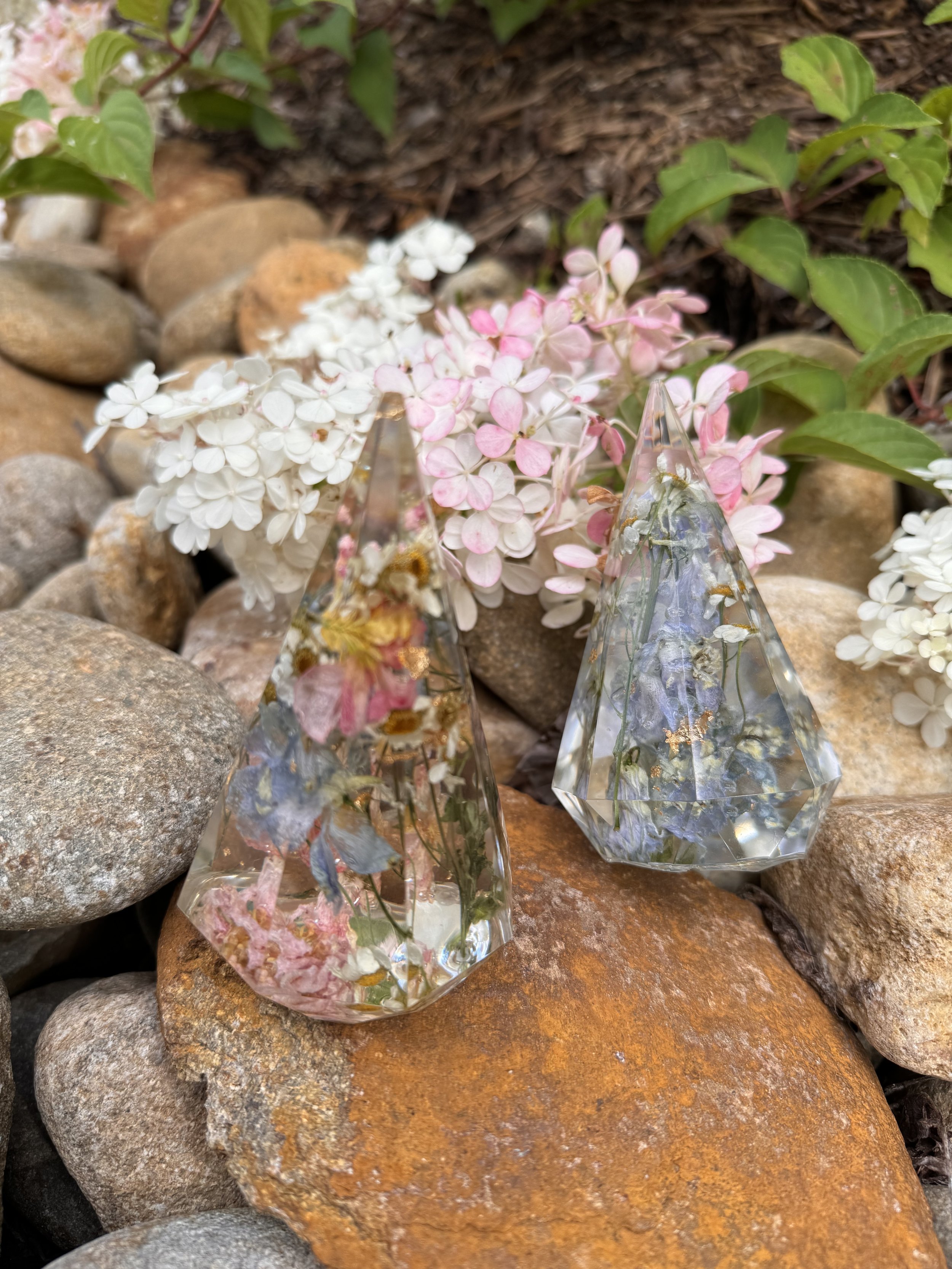 Two crystal pyramids containing dried flowers on rocks, surrounded by white and pink hydrangeas.