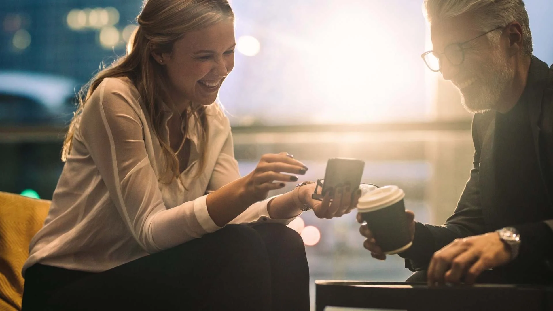 Une femme et un homme souriants discutant dans un café, assis l'un en face de l'autre, la femme montrant quelque chose sur son téléphone et tenant un café, la lumière du soleil en arrière-plan.