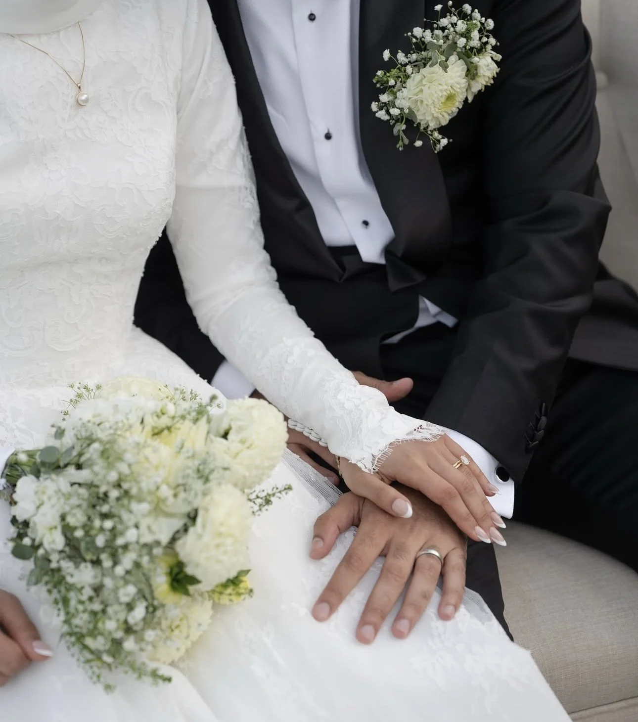 Close-up of a bride and groom holding hands, with wedding rings, both dressed in wedding attire, holding a bouquet of white flowers.