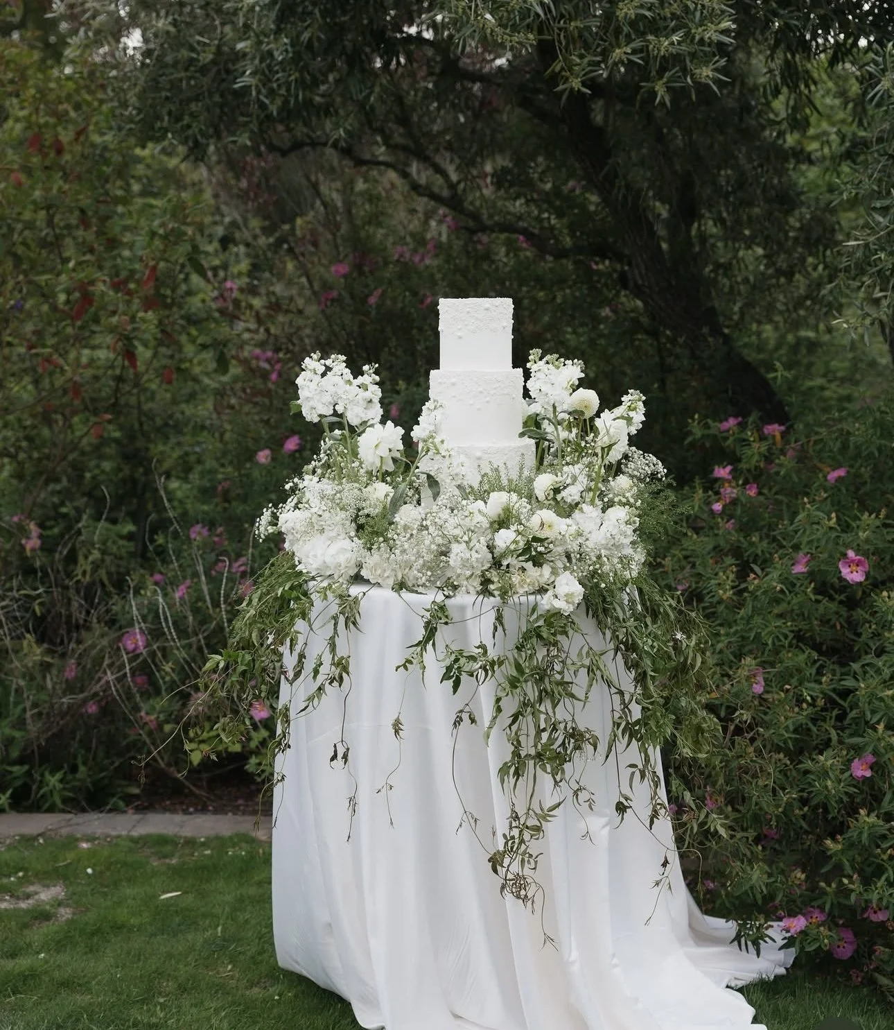 A white three-tier wedding cake decorated with white flowers, displayed on a table with white drapery and greenery, outdoors in a garden setting.