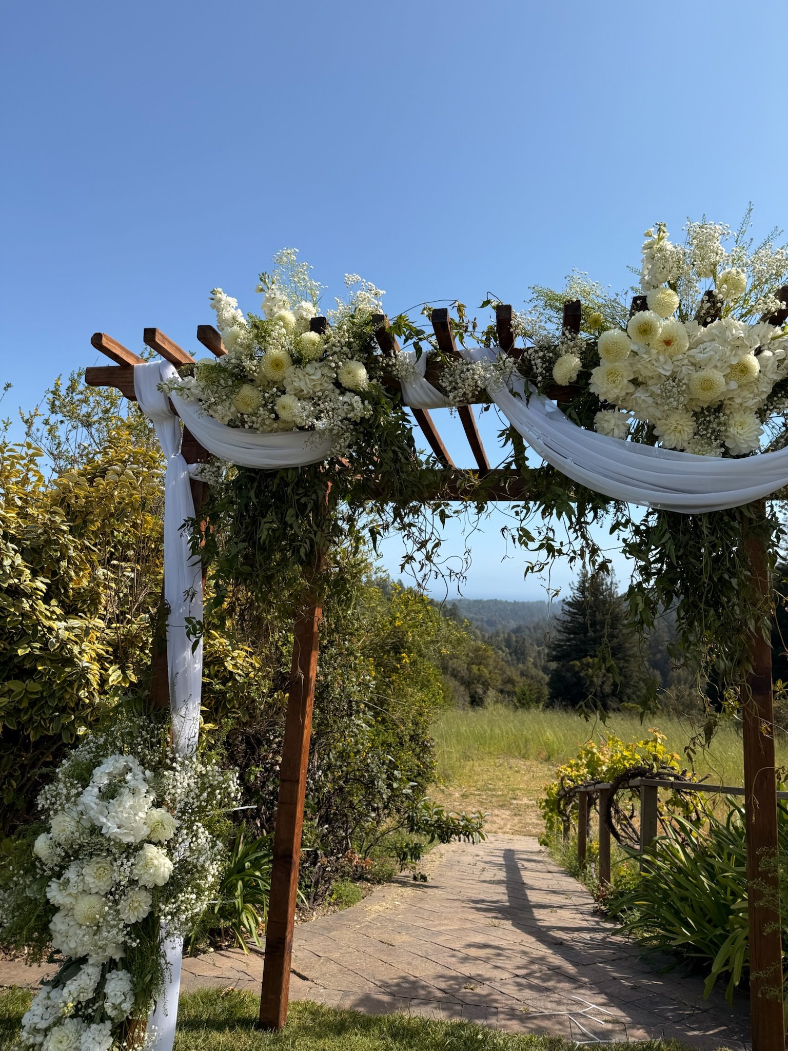 A decorated wedding arch with white flowers and white fabric drapes overlooking a garden pathway.
