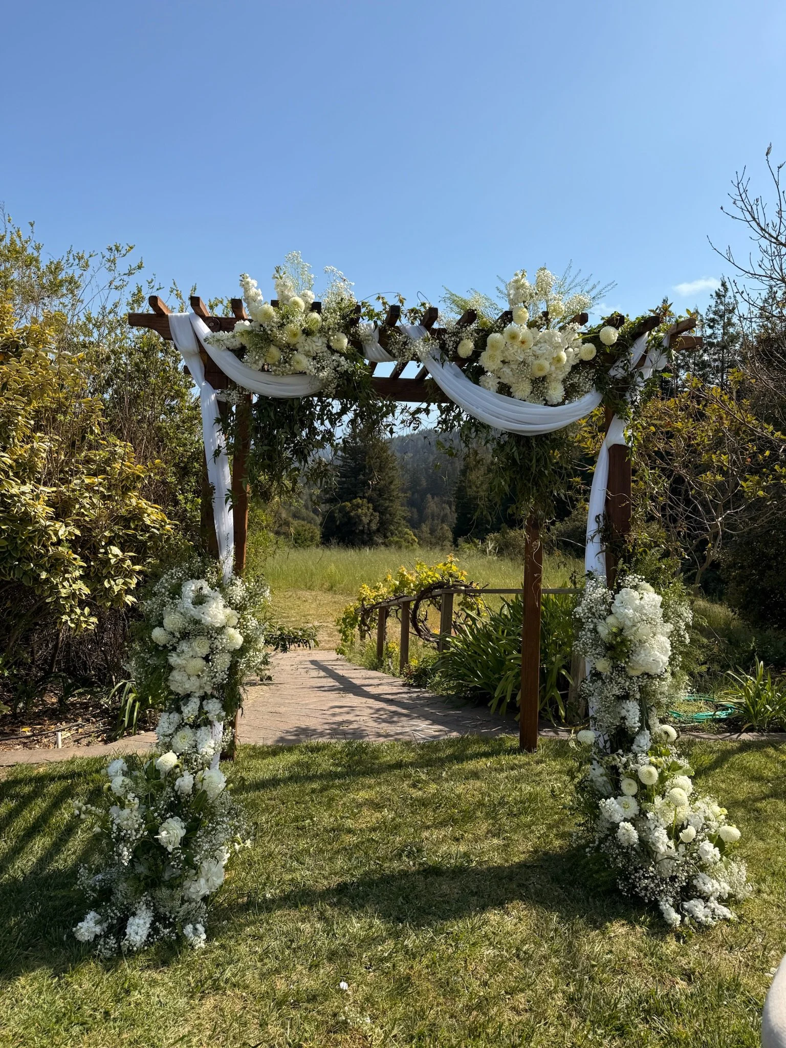 A decorated wedding arch with white flowers and draped white fabric set outdoors on grass with trees and clear blue sky in the background.