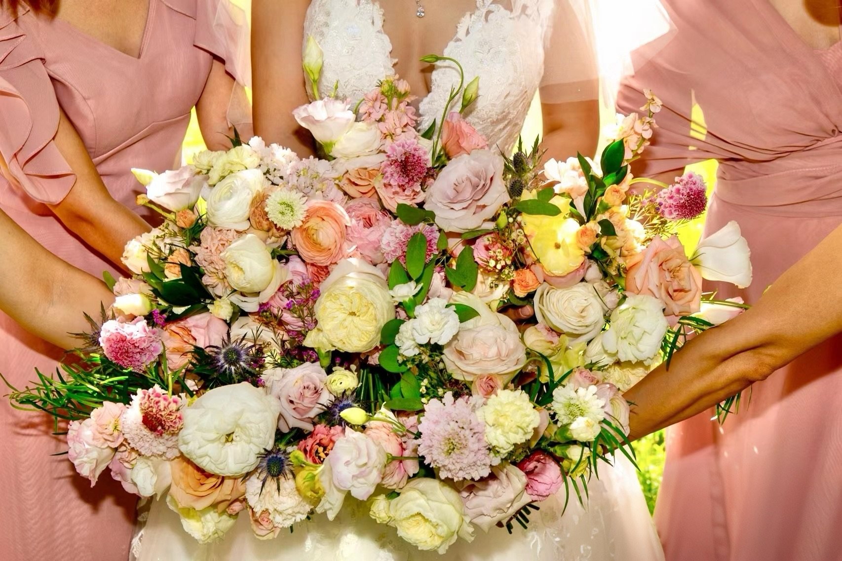 A bride holding a large bouquet of pink, white, and cream flowers, surrounded by two women in pink dresses, outdoors with sunlight.