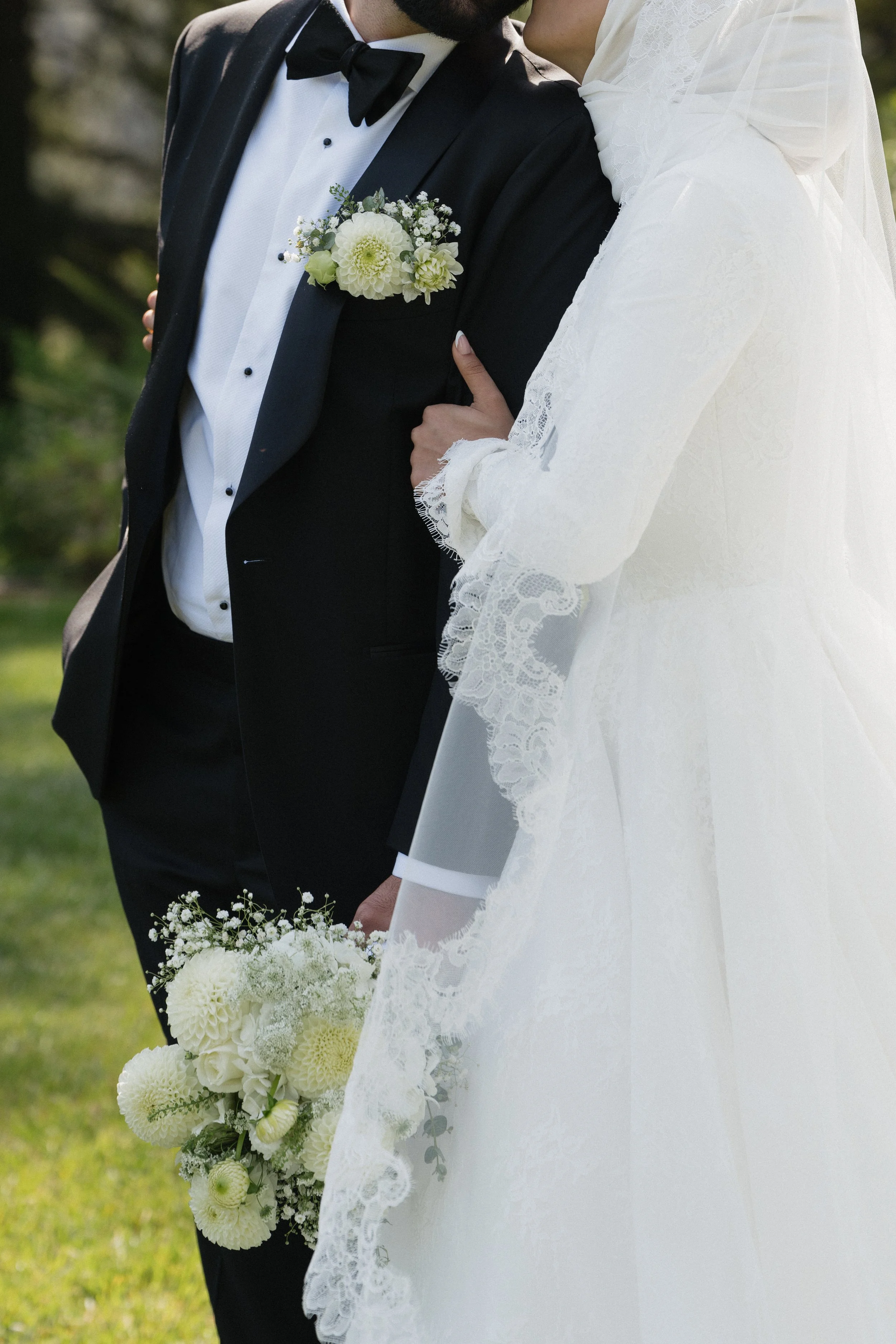 Close-up of a bride and groom at their wedding, with the bride in a white lace gown and the groom in a black tuxedo, holding a bouquet of white flowers.