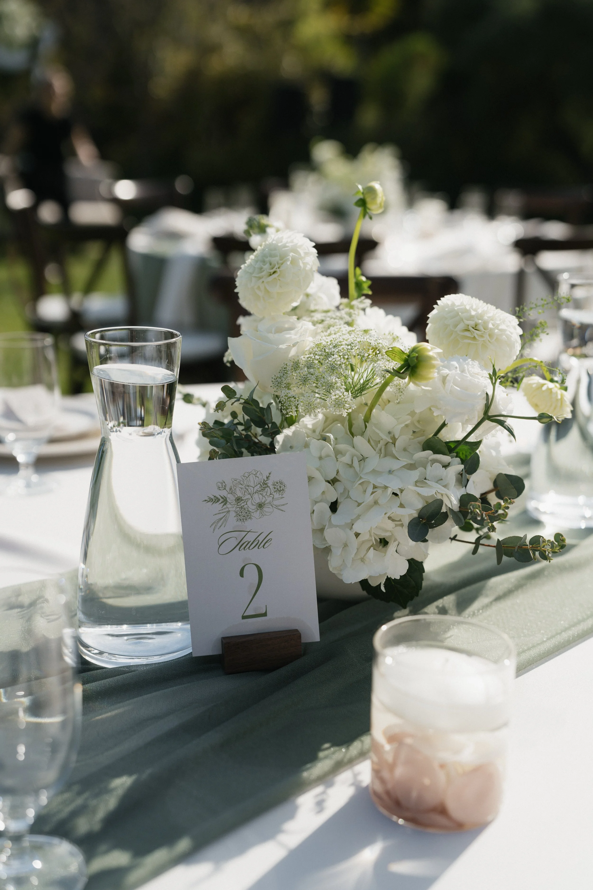 Elegant outdoor wedding reception table decorated with a floral centerpiece of white flowers, a water pitcher, drinking glasses, and a table number card.