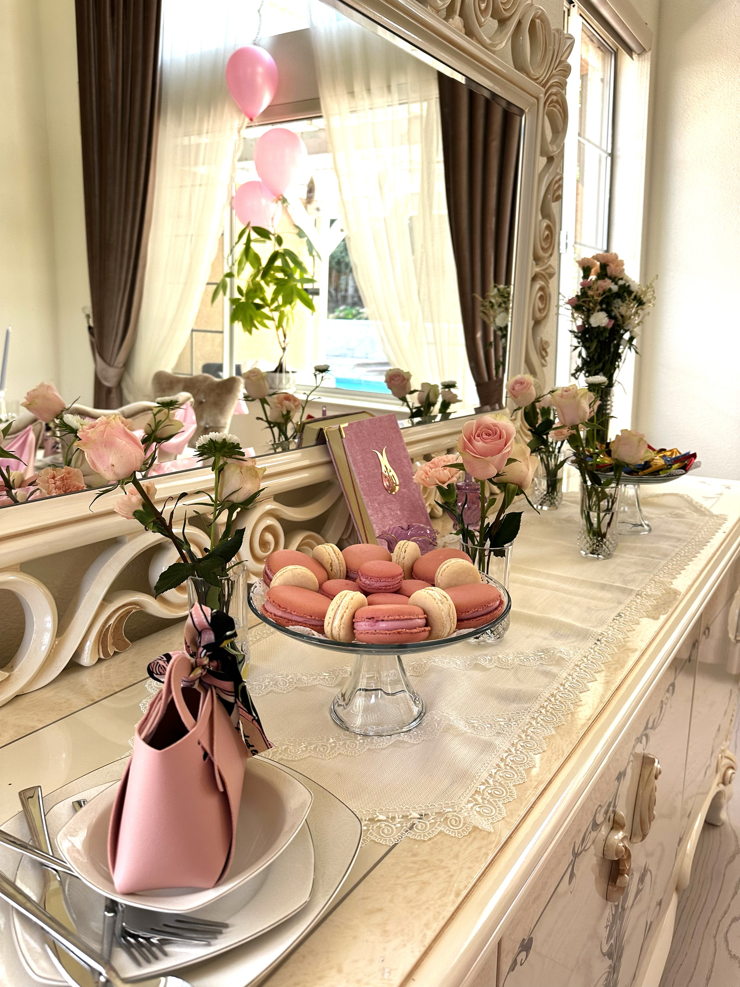 Decorative table with pink roses, macarons, and pink gift bag, reflected in large ornate mirror with cream curtains, pink balloons outside window, and additional flowers.