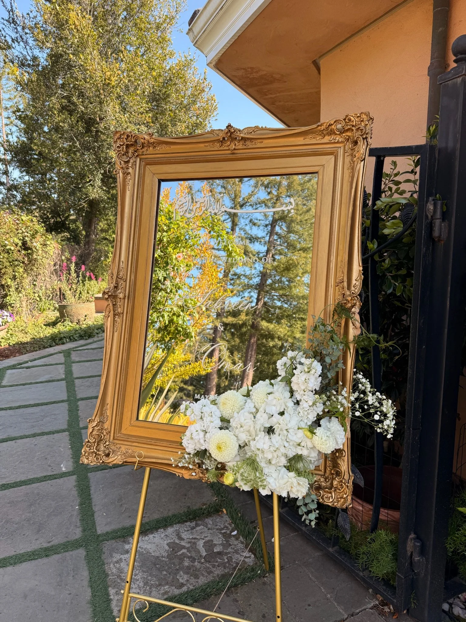 A decorative gold-framed mirror with engraved text, placed outdoors on a metal stand, decorated with a white floral arrangement, against a backdrop of trees and a garden area.