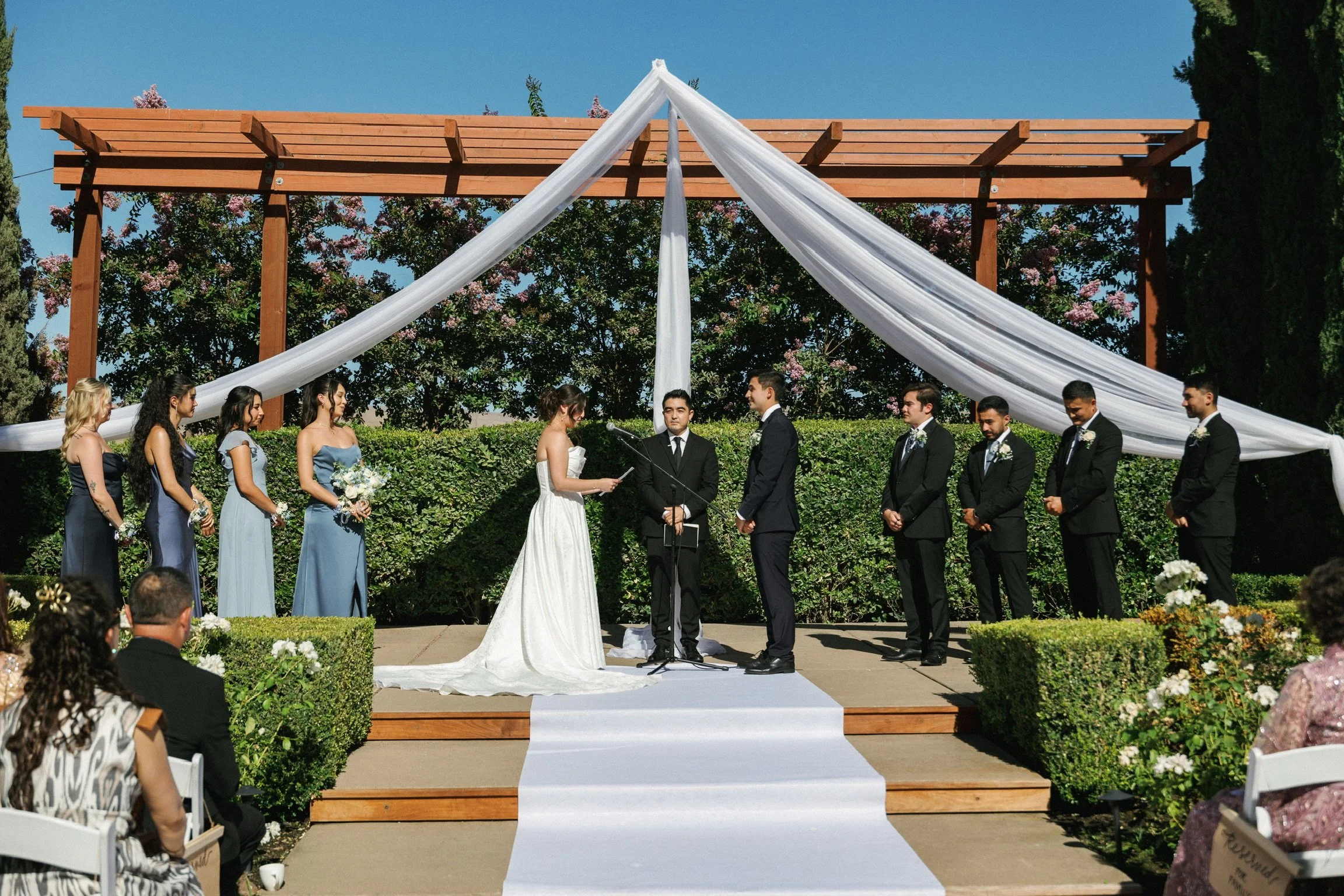 Outdoor wedding ceremony with bride and groom exchanging vows on a wooden platform decorated with white fabric, surrounded by bridesmaids and groomsmen in formal attire, with guests seated in front.