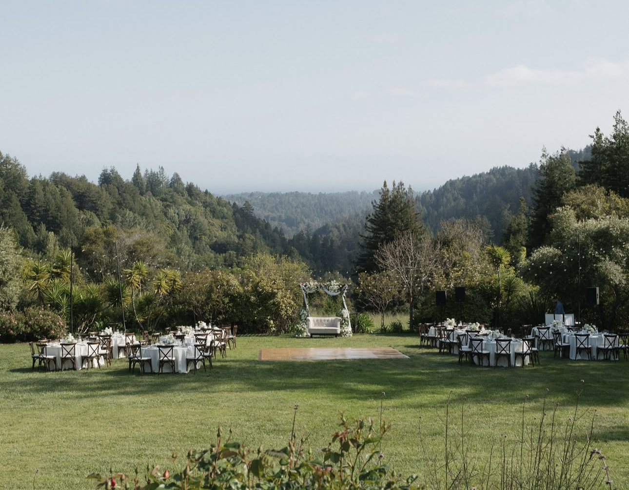 An outdoor wedding setup on a grassy field with round tables covered in white tablecloths and dark chairs, a decorated archway for the ceremony, and a scenic backdrop of green hills and trees under a clear sky.