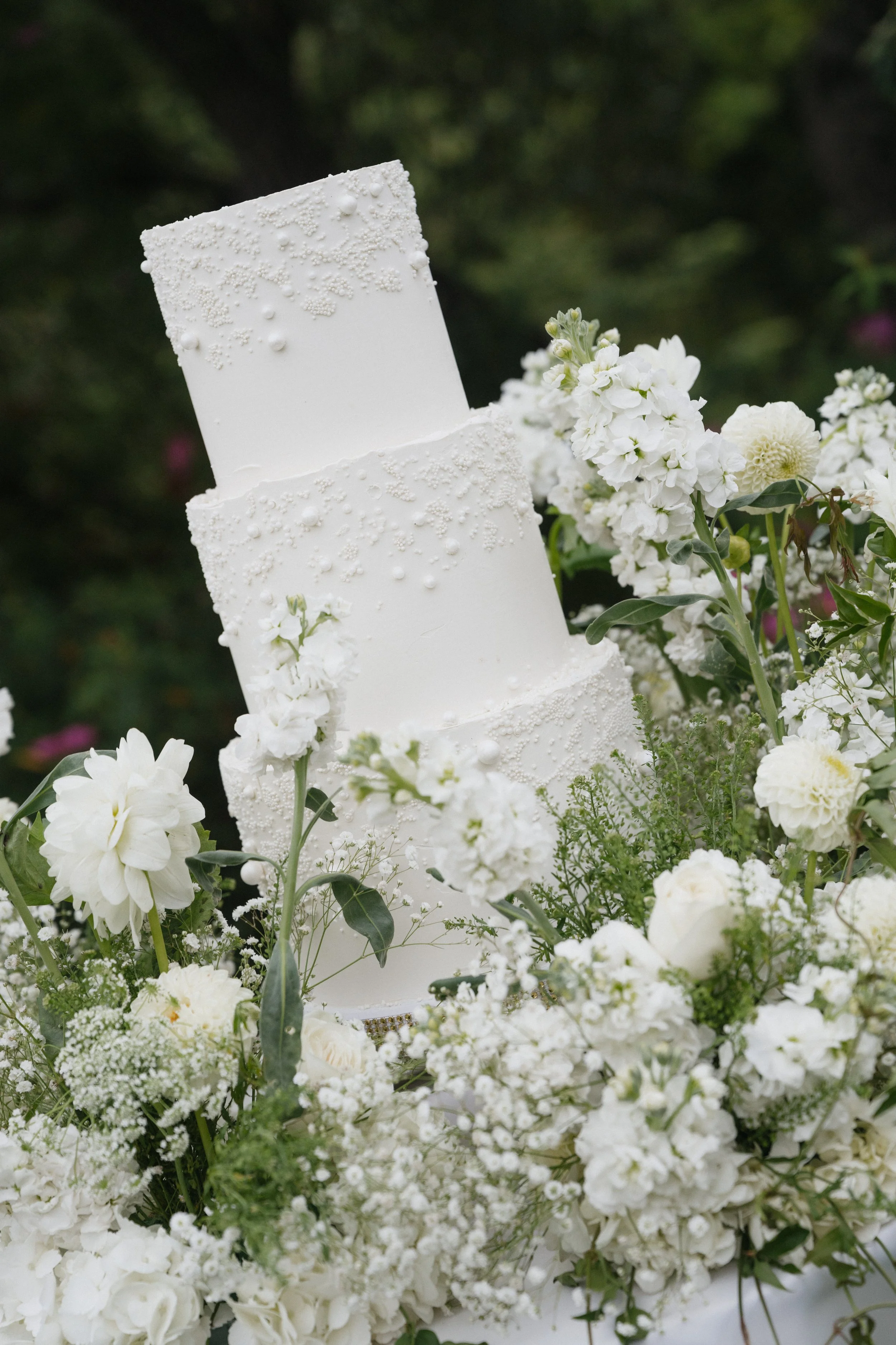 A white, three-tier wedding cake decorated with small white beads and sugar pearls, surrounded by white flowers and greenery.
