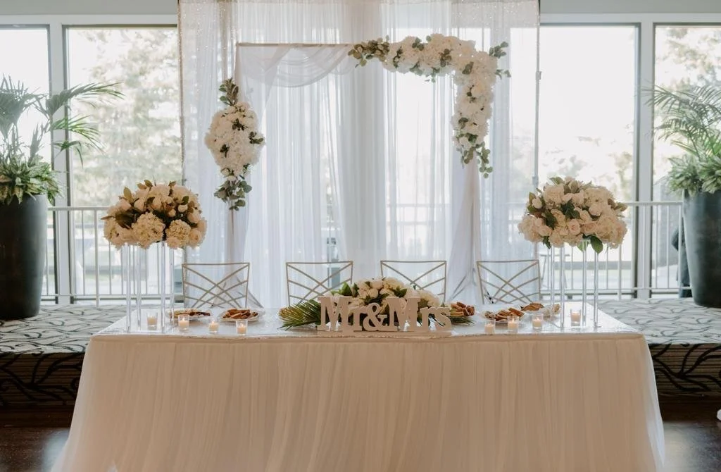 Wedding reception table with floral arrangements and a white draped backdrop, labeled 'Mr. & Mrs.'