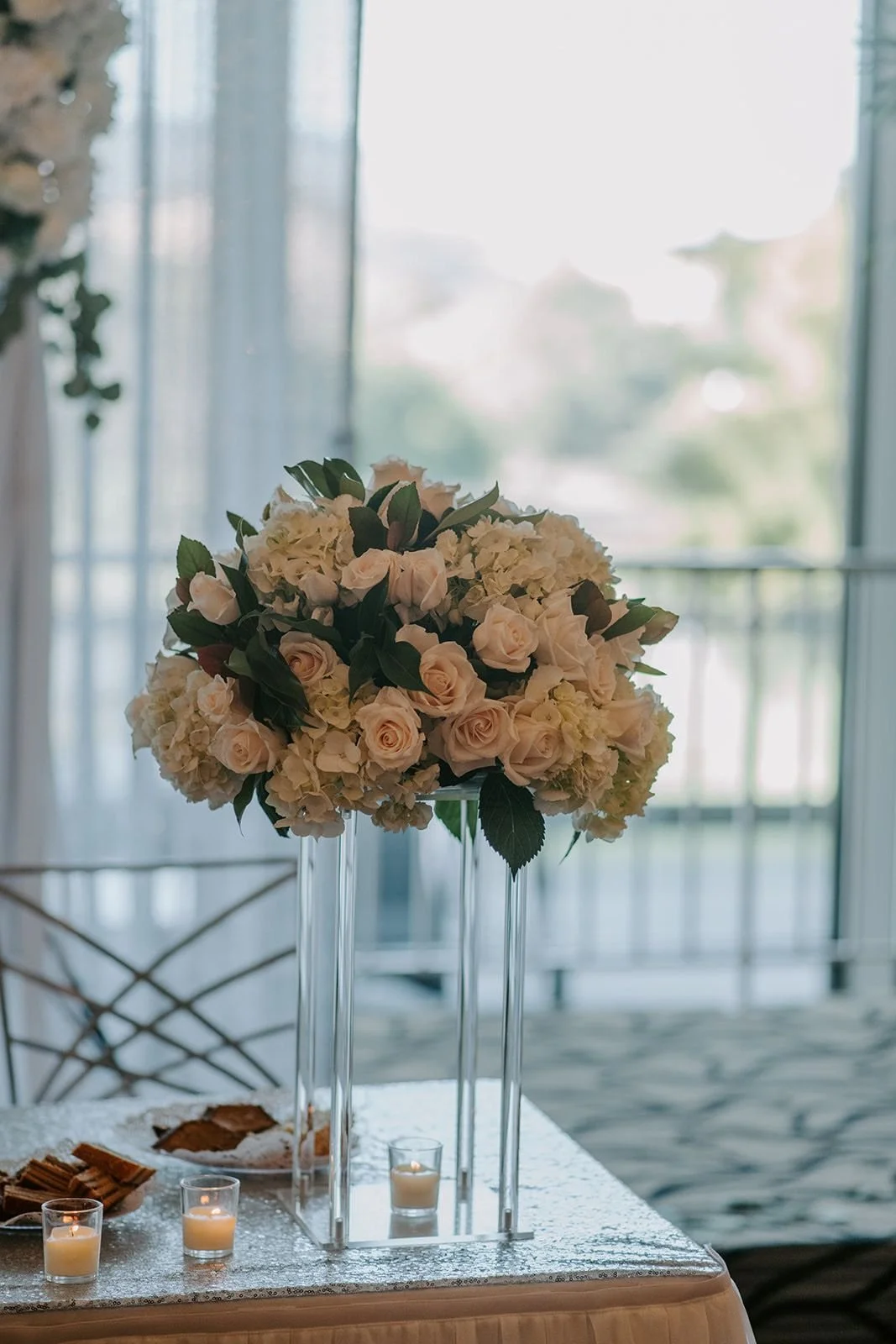 Elegant floral centerpiece made of white roses and hydrangeas on a tall glass stand, placed on a table with candles and snacks, with a background of sheer curtains and a balcony door.