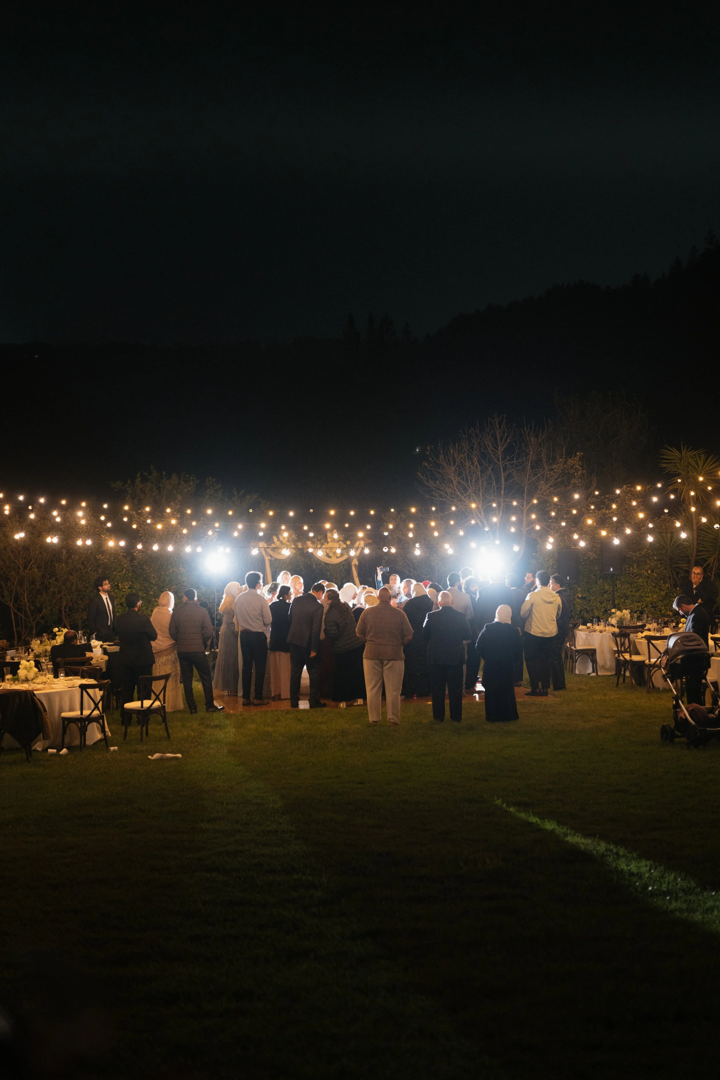 Nighttime outdoor wedding reception with string lights, people gathered, some dancing and socializing, tables with white tablecloths and floral centerpieces, surrounded by trees and hills in the background.