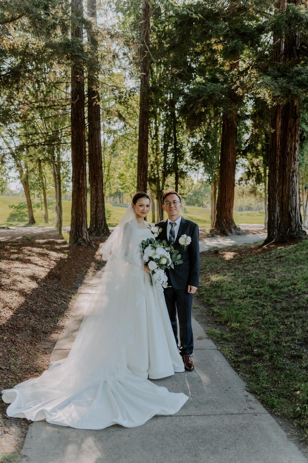 A bride and groom standing on a sidewalk in a park with tall trees, the bride wearing a white wedding gown and veil holding a bouquet, the groom in a dark suit, both smiling.