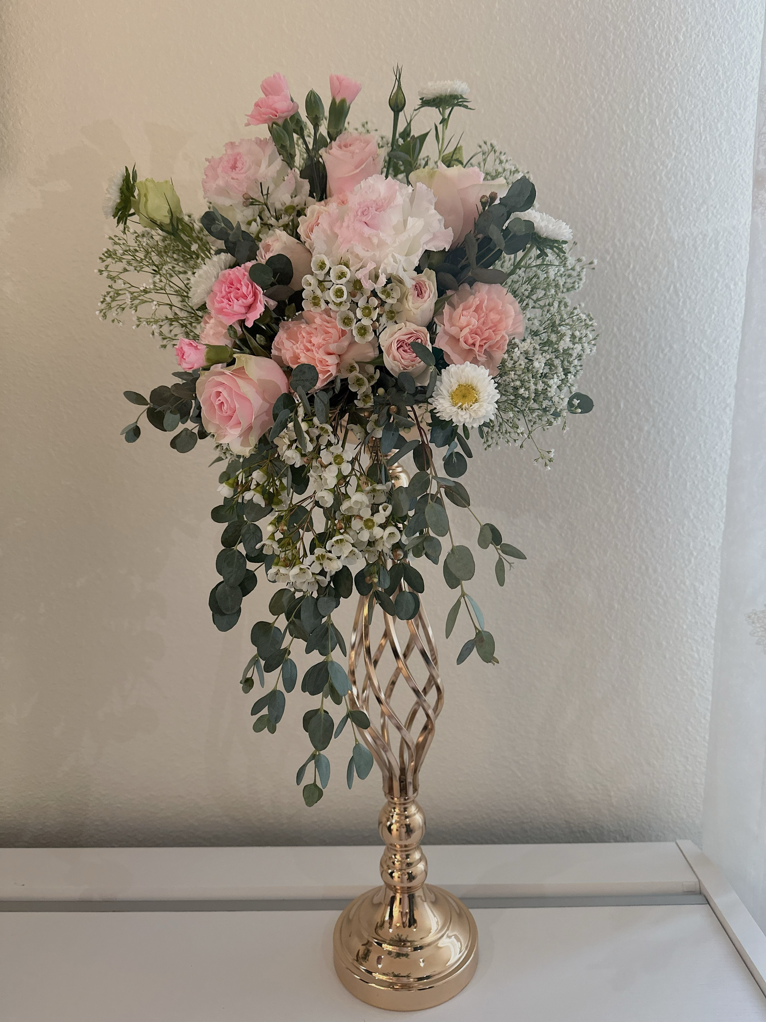 A tall, ornate gold-colored vase holding a bouquet of pink and white flowers, including roses, daisies, and greenery, against a beige wall.