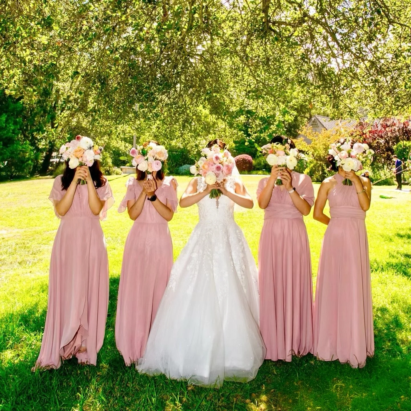 Bride and four bridesmaids posing outdoors in a green garden, holding bouquets in front of their faces, with trees and sunlight in the background.