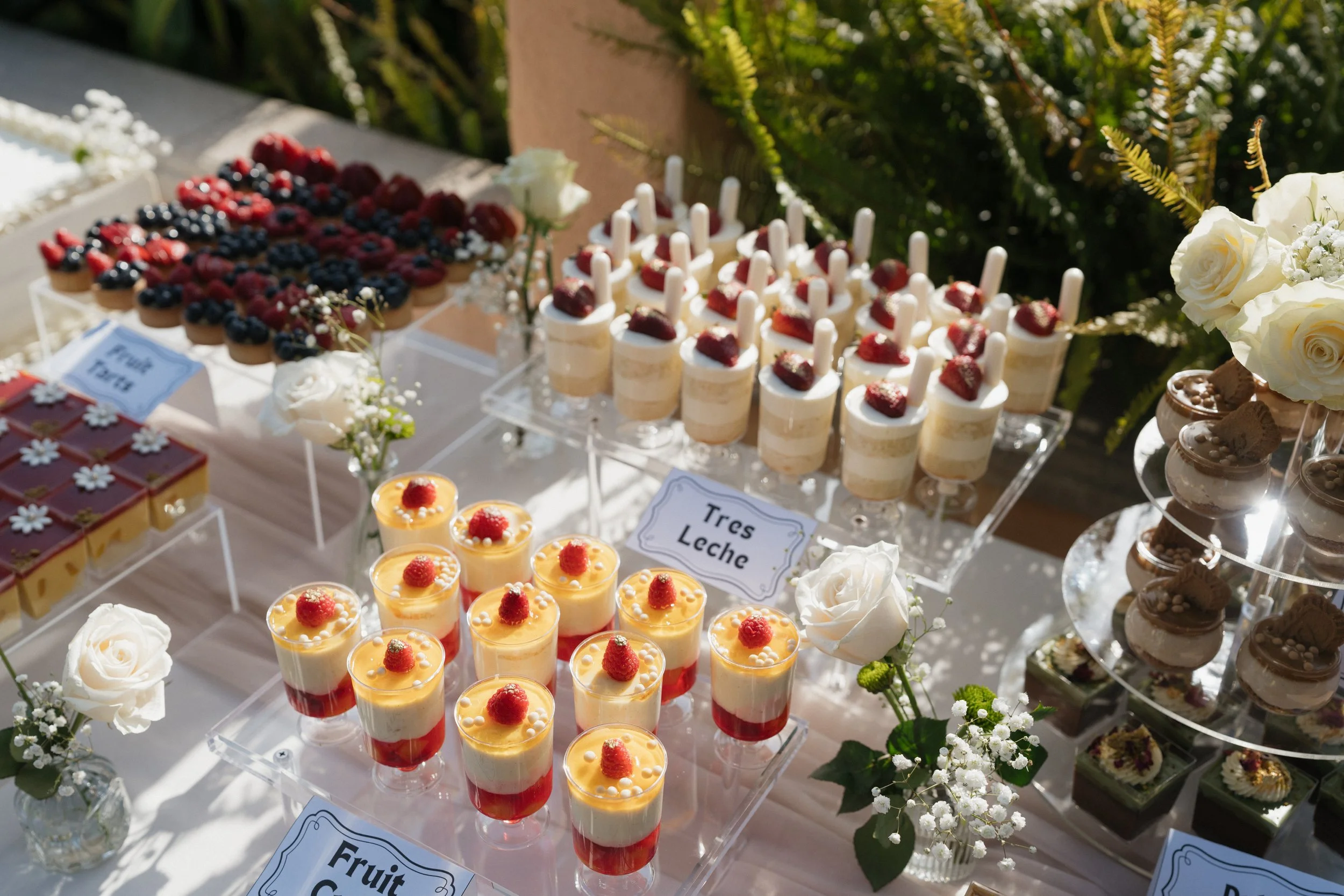 Dessert table with various fruit and cream desserts, decorated with white roses and small white flowers, outdoors in sunlight.