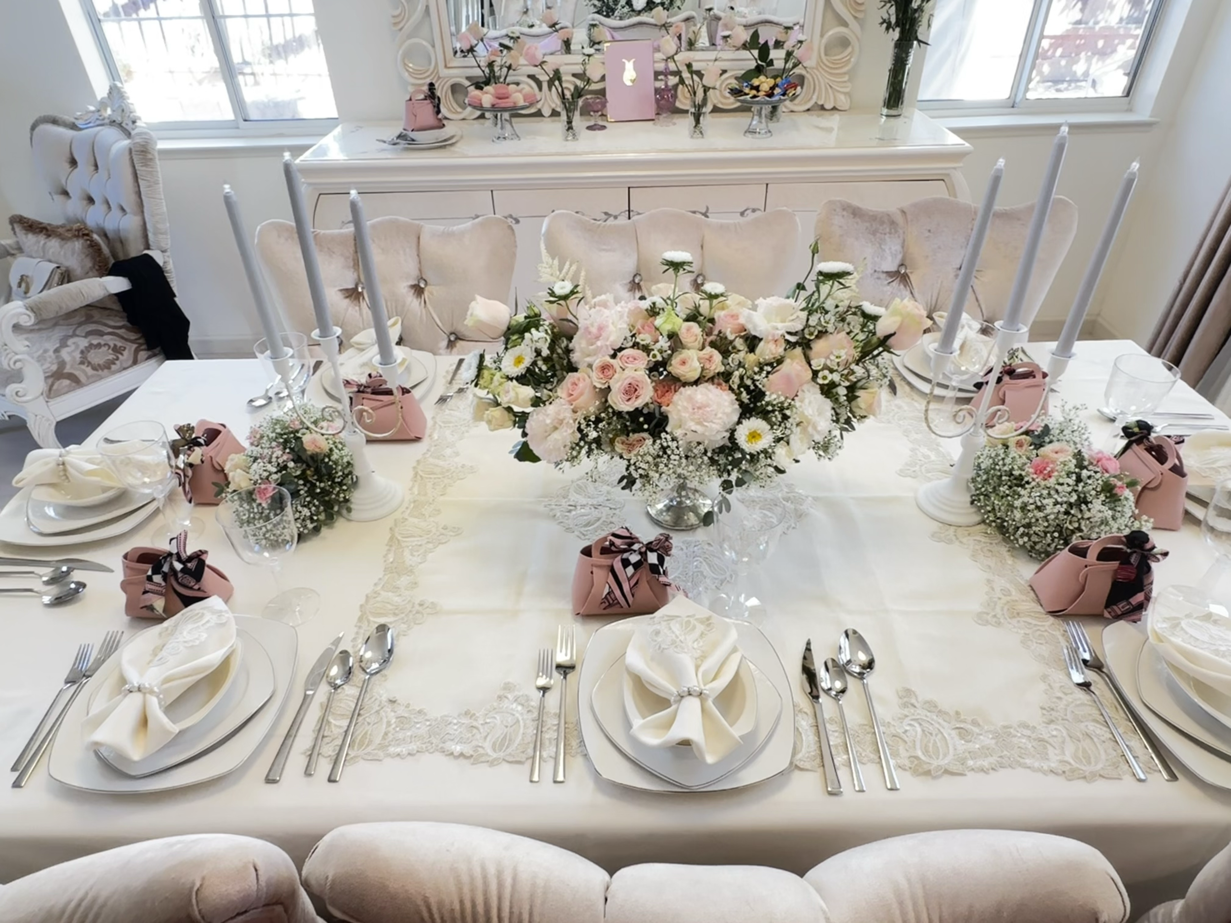 Elegant dining table decorated with a large central bouquet of pink and white roses, surrounded by smaller floral arrangements and pink wrapped gifts, set with white plates, silverware, napkins, and tall white candlesticks in an airy, bright room wit
