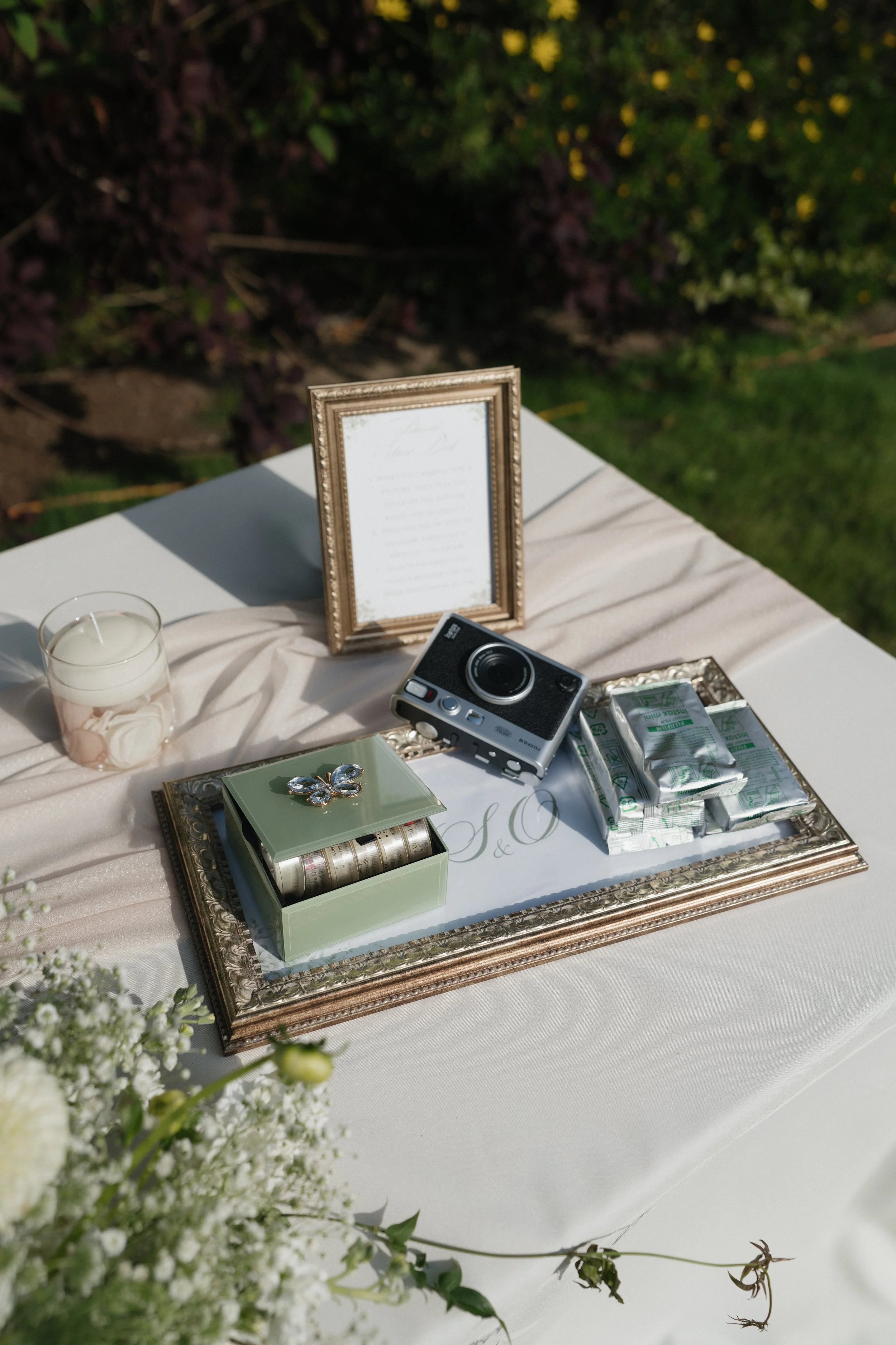 Decorative table setting with framed menu, camera, candle, and jewelry, outdoors with greenery.