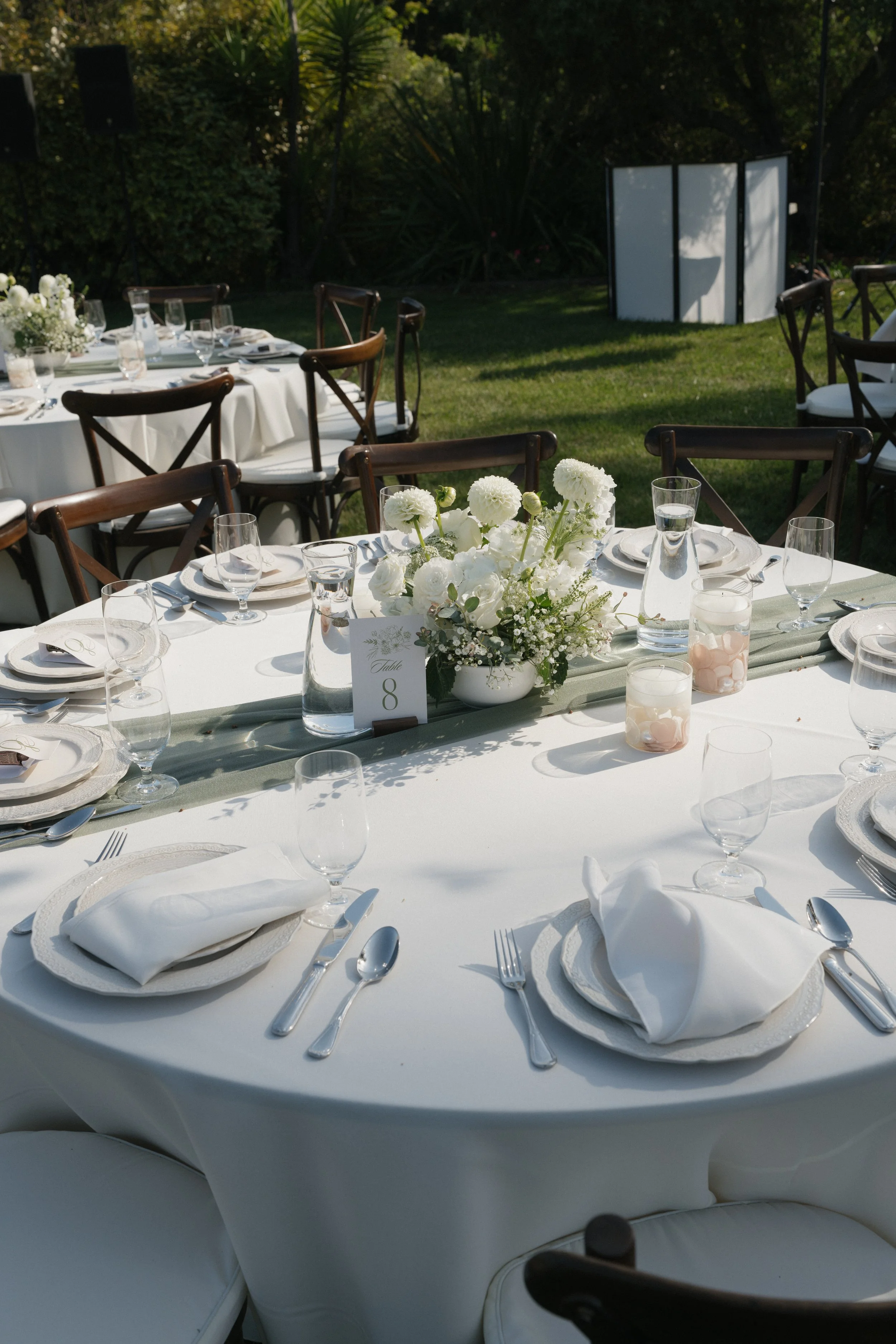 Elegant outdoor banquet table set with white tablecloths, white napkins, glassware, silverware, and floral centerpieces featuring white flowers on a green table runner, in a garden setting.