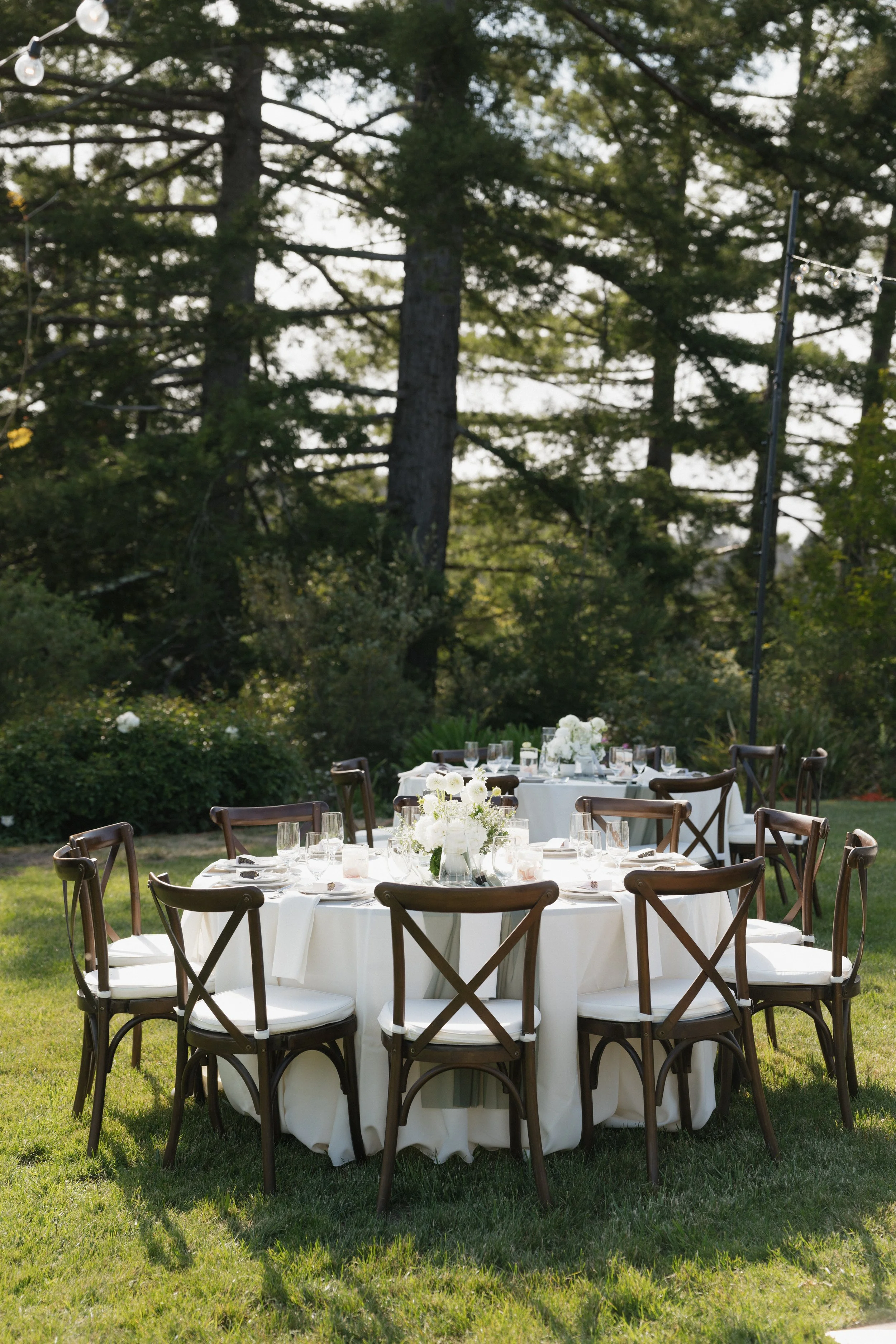 Round outdoor table with white tablecloth, decorated with white floral centerpieces, surrounded by wooden chairs with white cushions, set for a formal event in a grassy area with trees in the background.