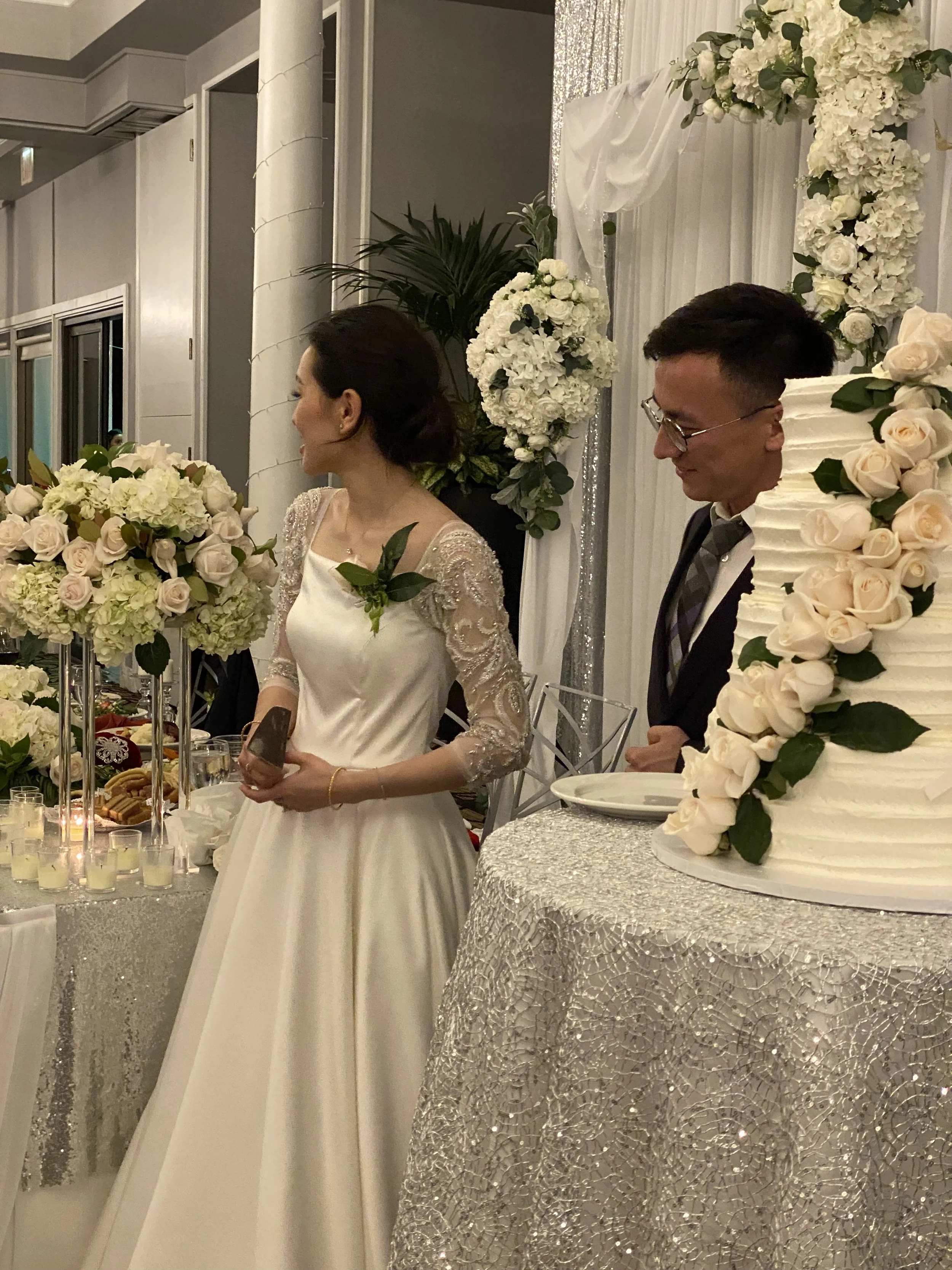 A bride and groom at a wedding reception standing beside a large decorated wedding cake surrounded by floral arrangements.