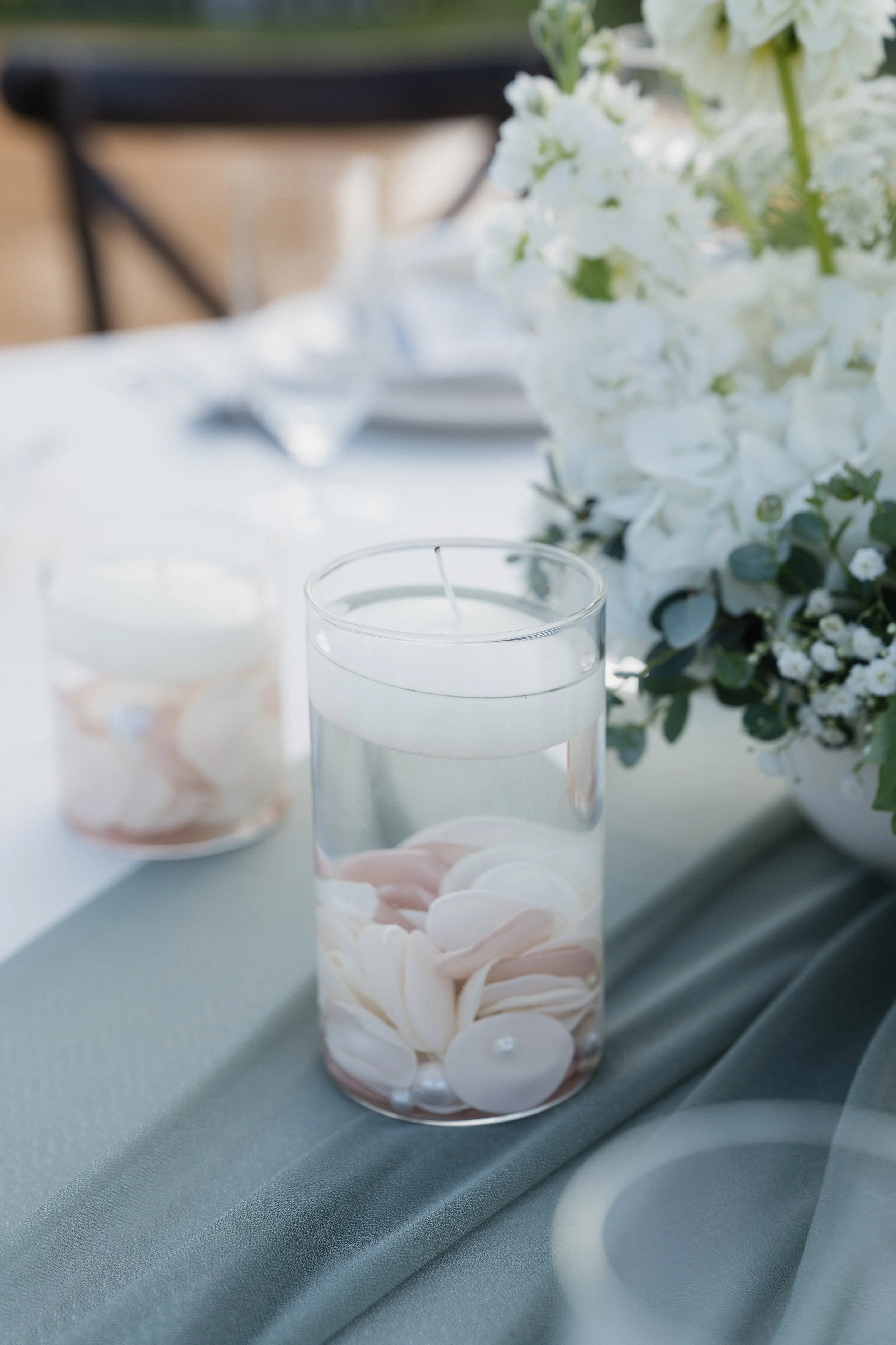 Vase with floating candles and pastel shell-shaped candles on a table with a floral centerpiece, likely at a wedding or special event.