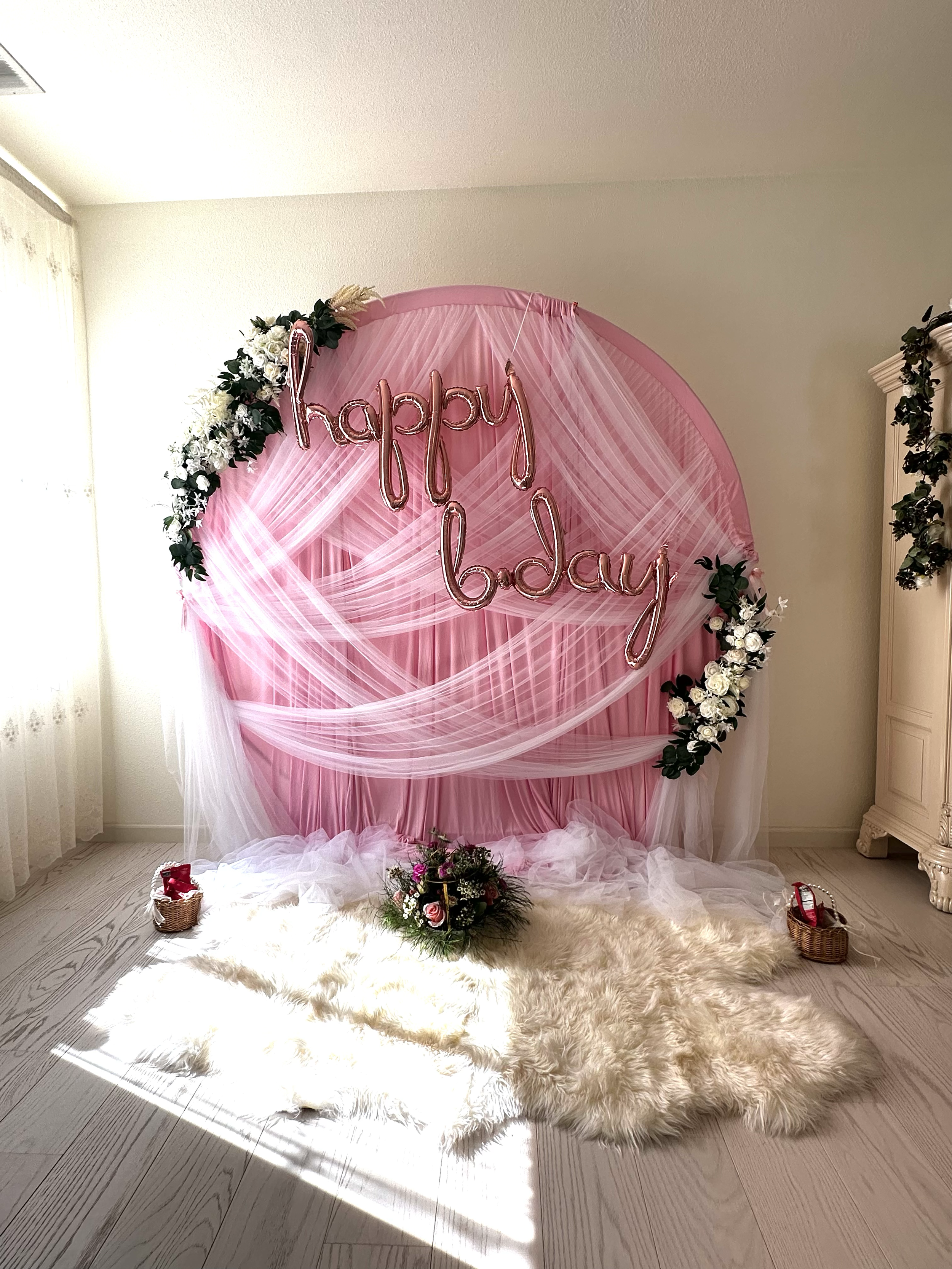 Decorative pink backdrop with white tulle drapes and a 'happy bday' balloon sign, floral arrangements, and baskets on a fluffy white rug in a bright room.