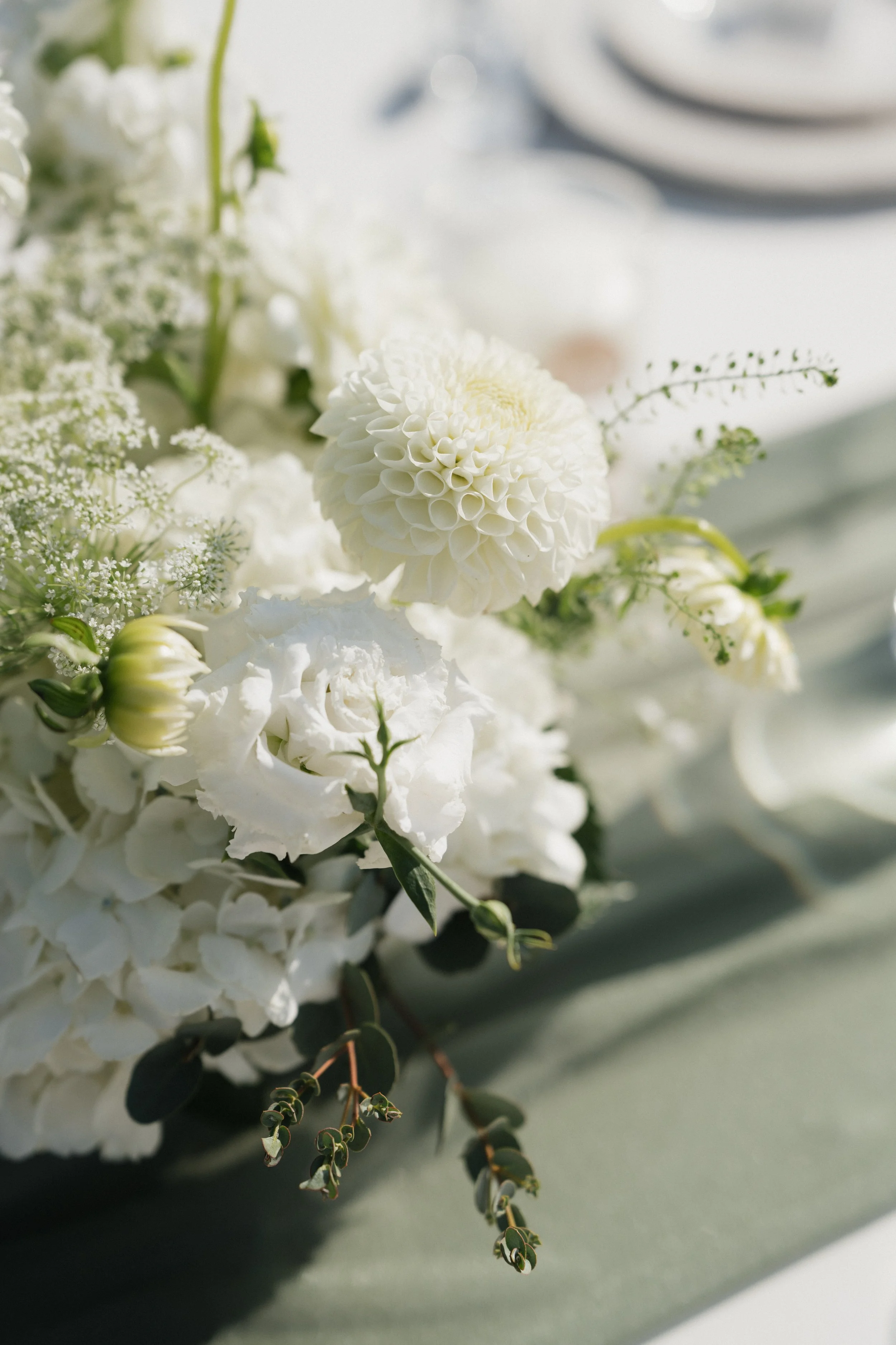 Close-up of white floral arrangement with dahlias, roses, and greenery on a table.