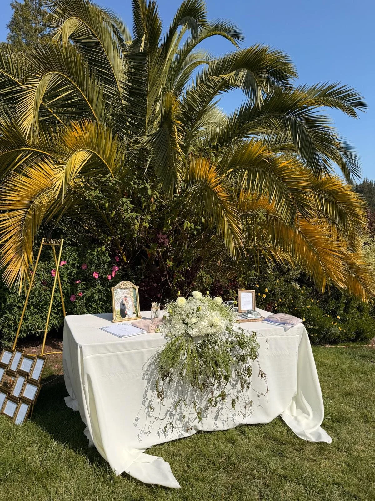 A decorated wedding sign-in table with a white tablecloth, floral arrangements, framed photographs, and picture frames, set outdoors in front of large palm trees and greenery.